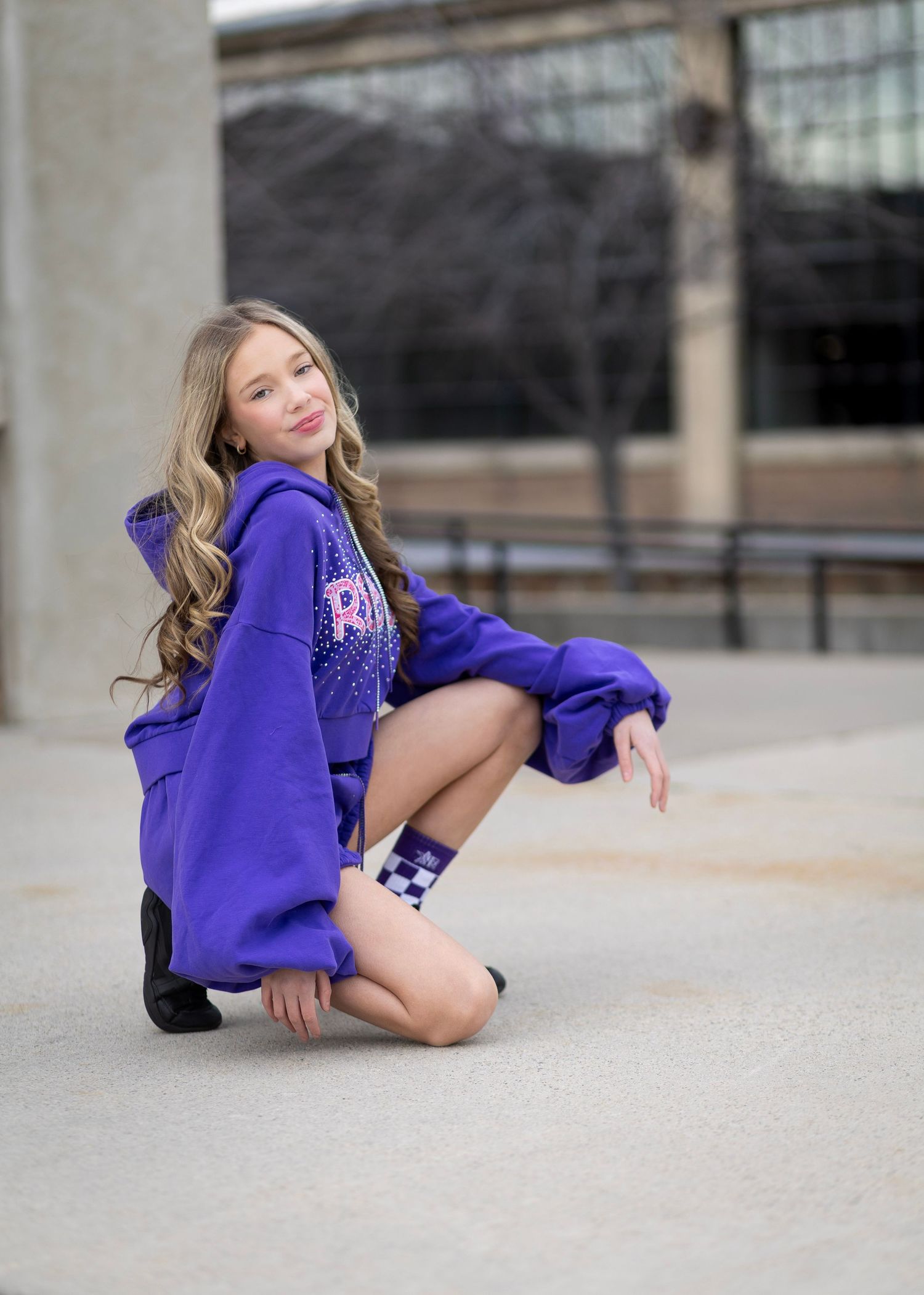 Young girl in purple hoodie and checkered socks poses outdoors on pavement in a candid fashion photo.