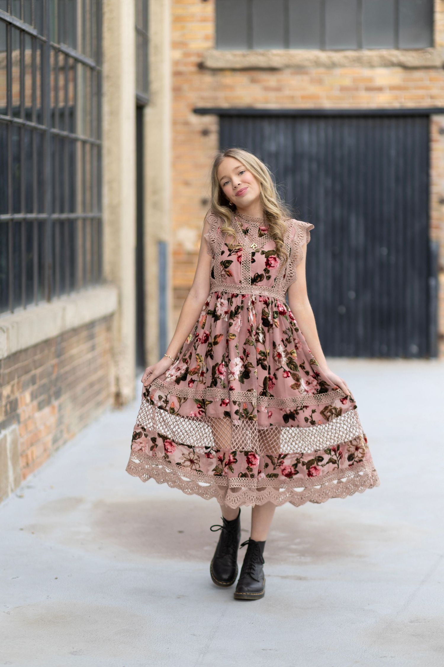 A girl twirls in a floral lace midi dress with black boots in an urban alleyway setting.