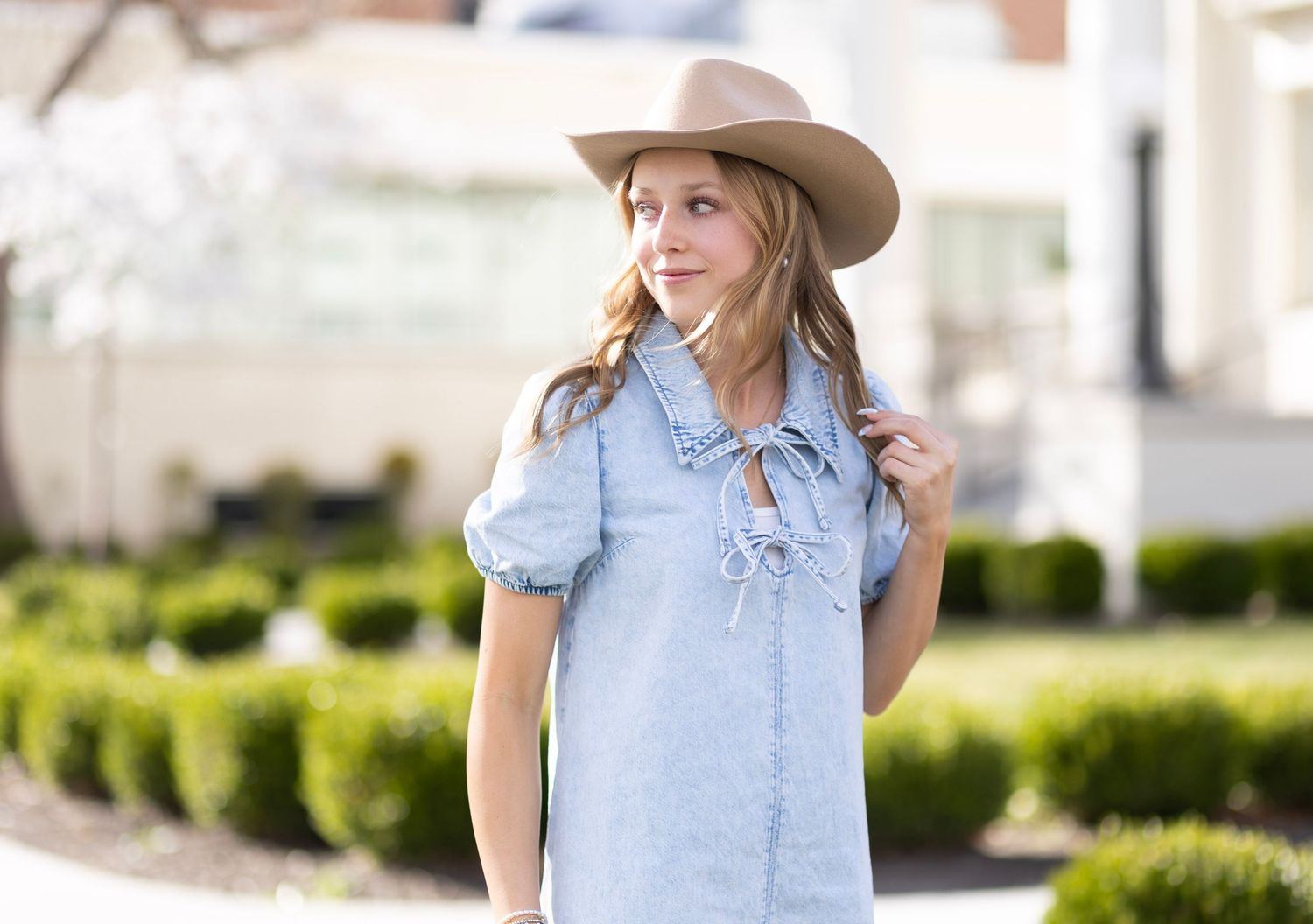 Young woman in a light blue chambray dress with puff sleeves and a tan cowboy hat poses outdoors.