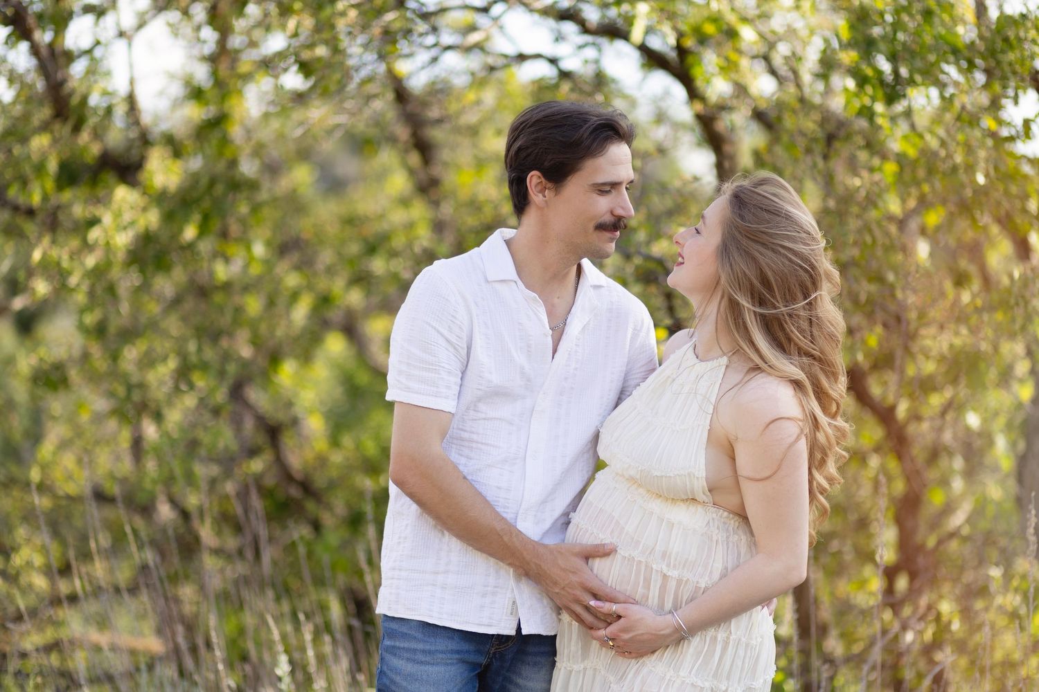 Happy couple embracing outdoors, pregnant woman in yellow dress with partner in white shirt among green trees.