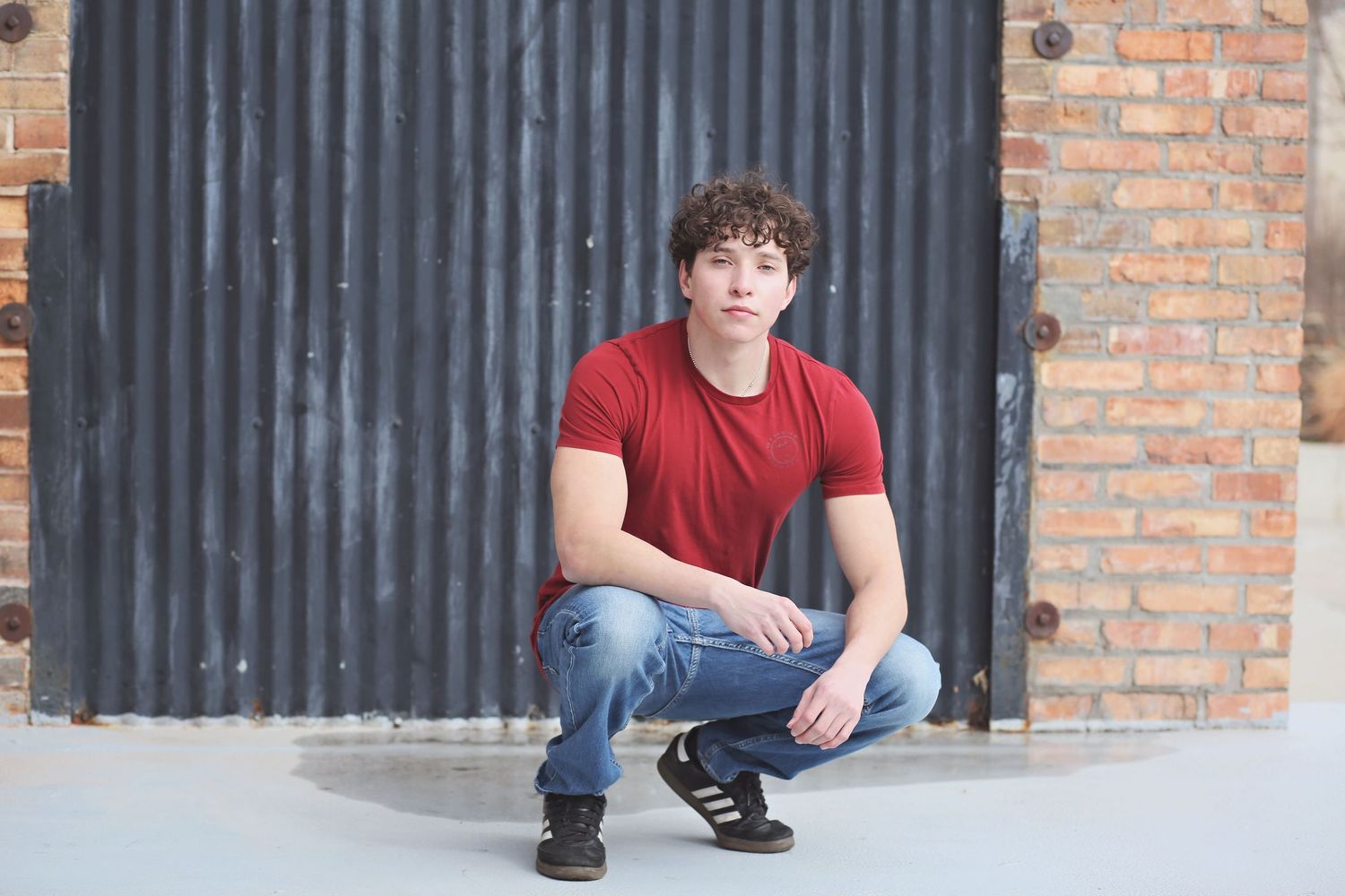 Person in red shirt and jeans crouching against industrial brick and metal wall.