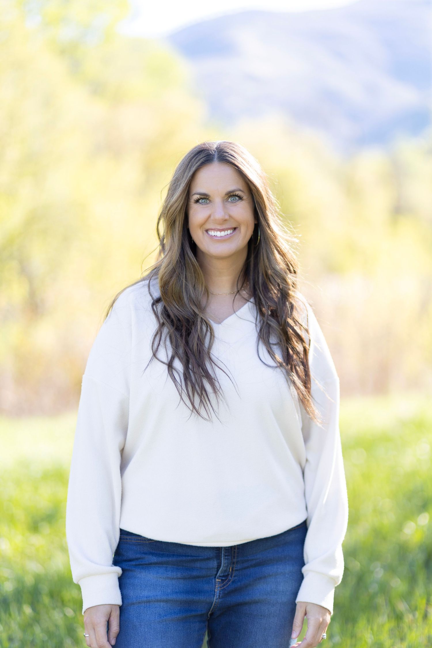 Smiling woman with long brown hair wearing a cream sweater and jeans standing outdoors with mountains behind her.
