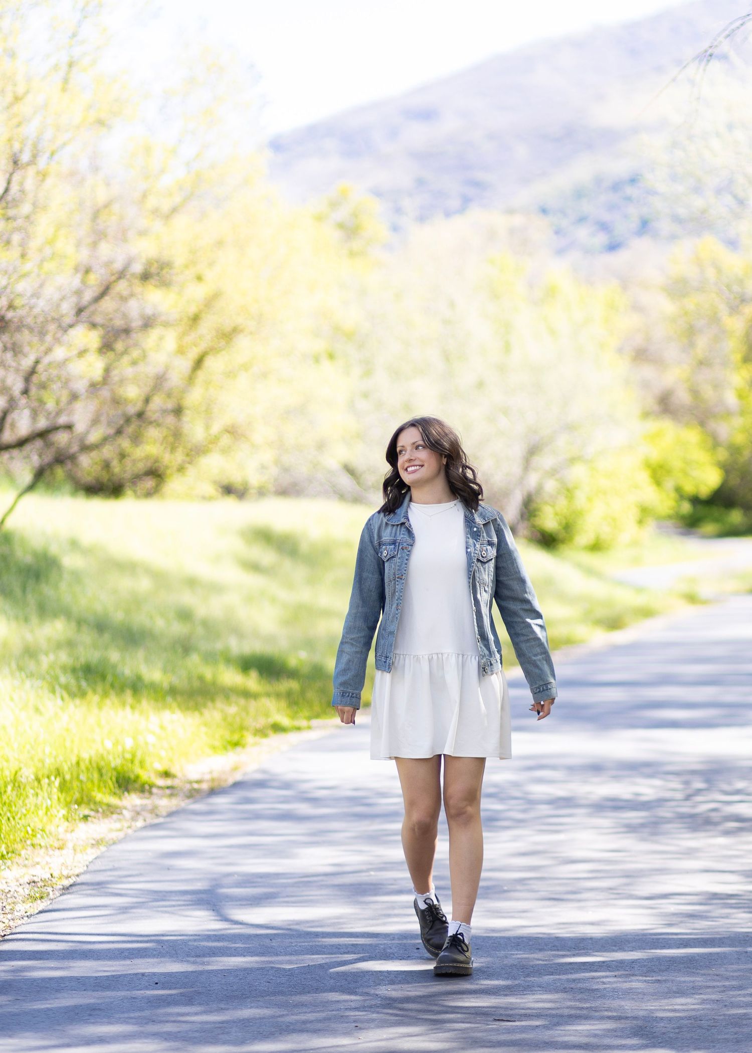 Teen girl in white dress and denim jacket walks along scenic tree-lined path with mountain view.