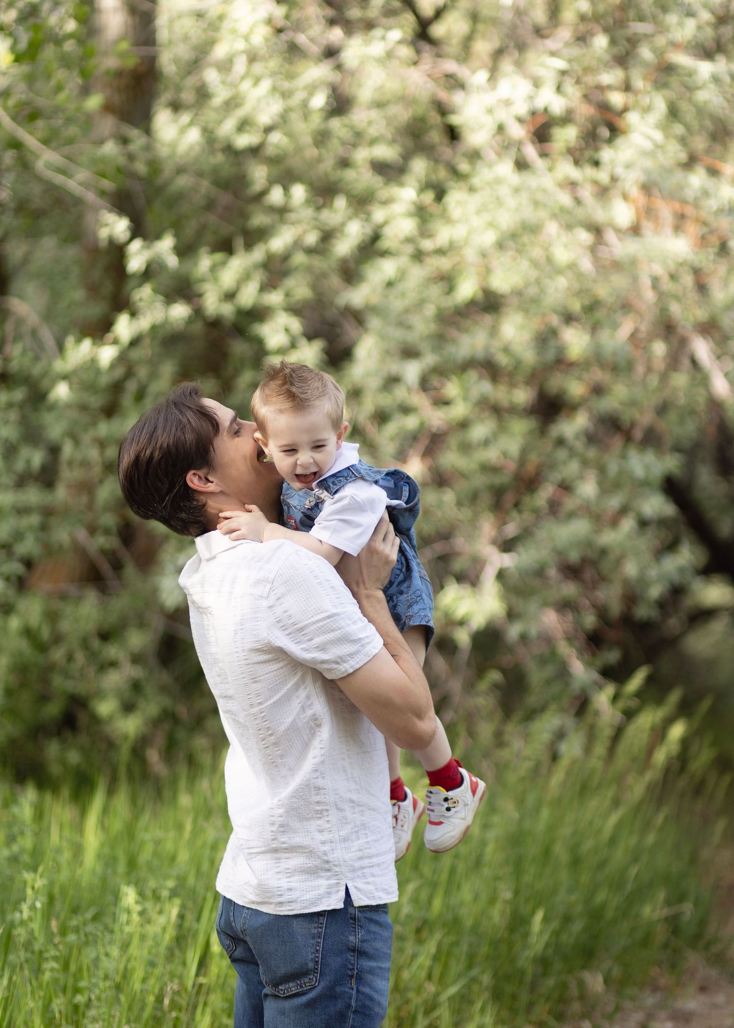 Parent kissing a happy toddler while holding them up outdoors in a lush green park setting.