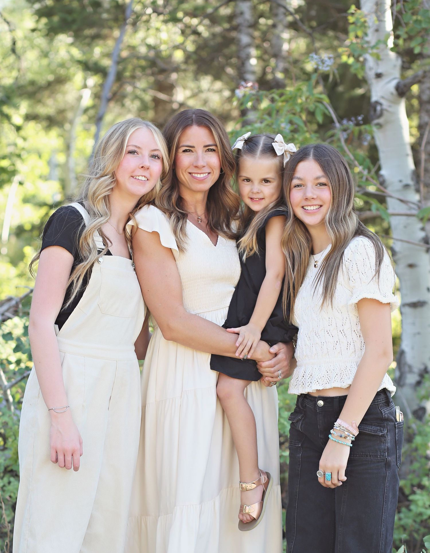 Four women in coordinating white and black outfits pose together outdoors among aspen trees.