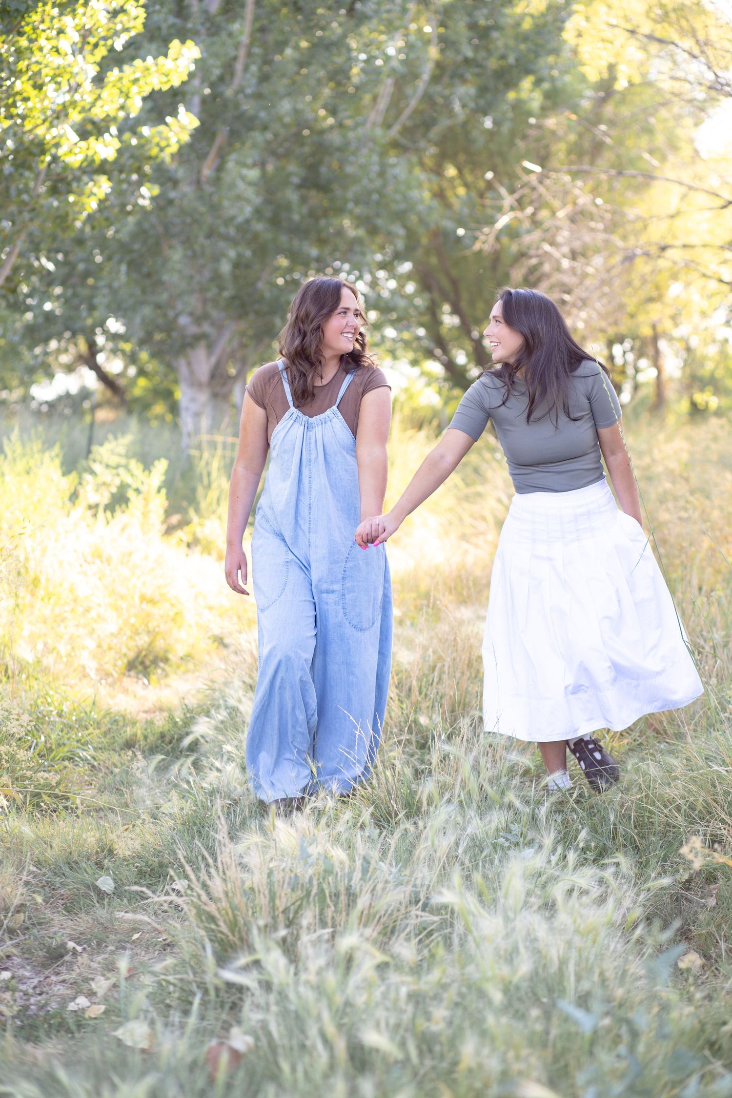Two people in casual denim outfits walk together through tall grass in sunlit meadow.
