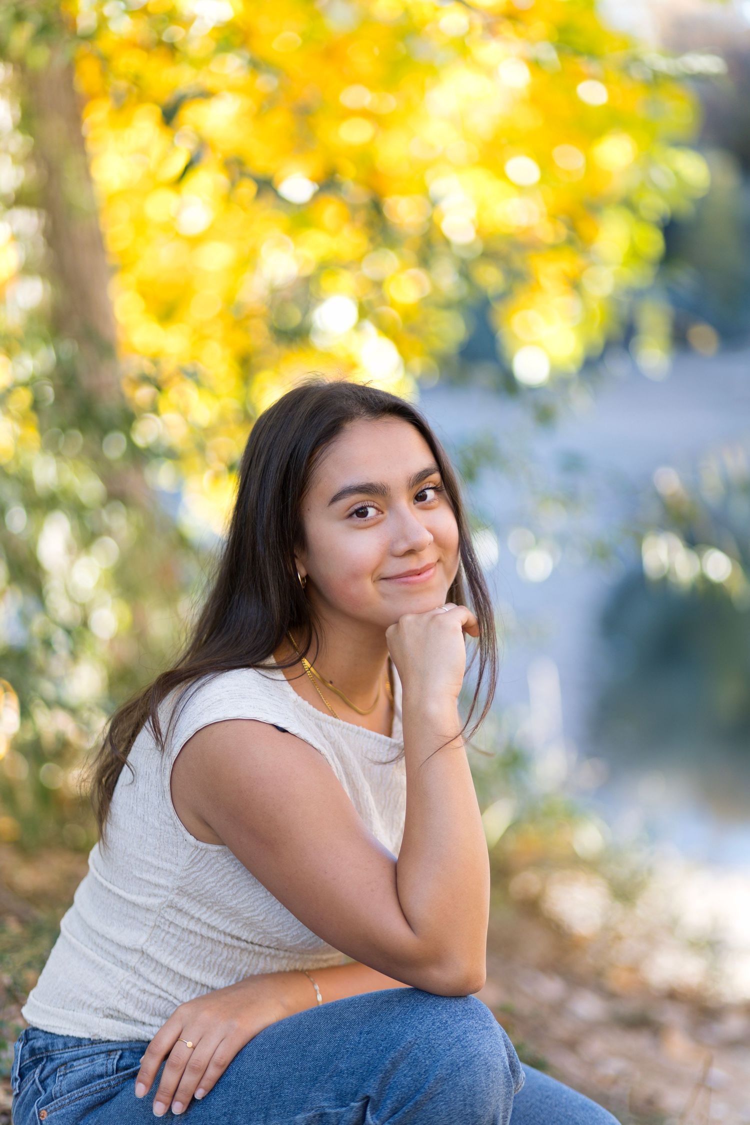 Young person in white sleeveless top and jeans sits thoughtfully outdoors with autumn foliage in background.