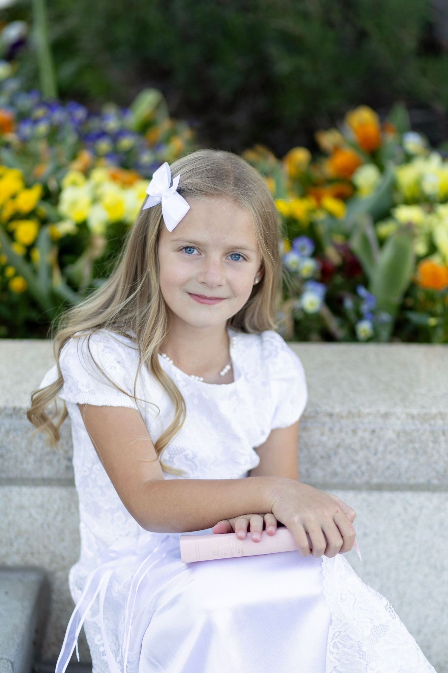 Young girl in white dress with bow holds pink book, sitting outdoors near colorful flowers.