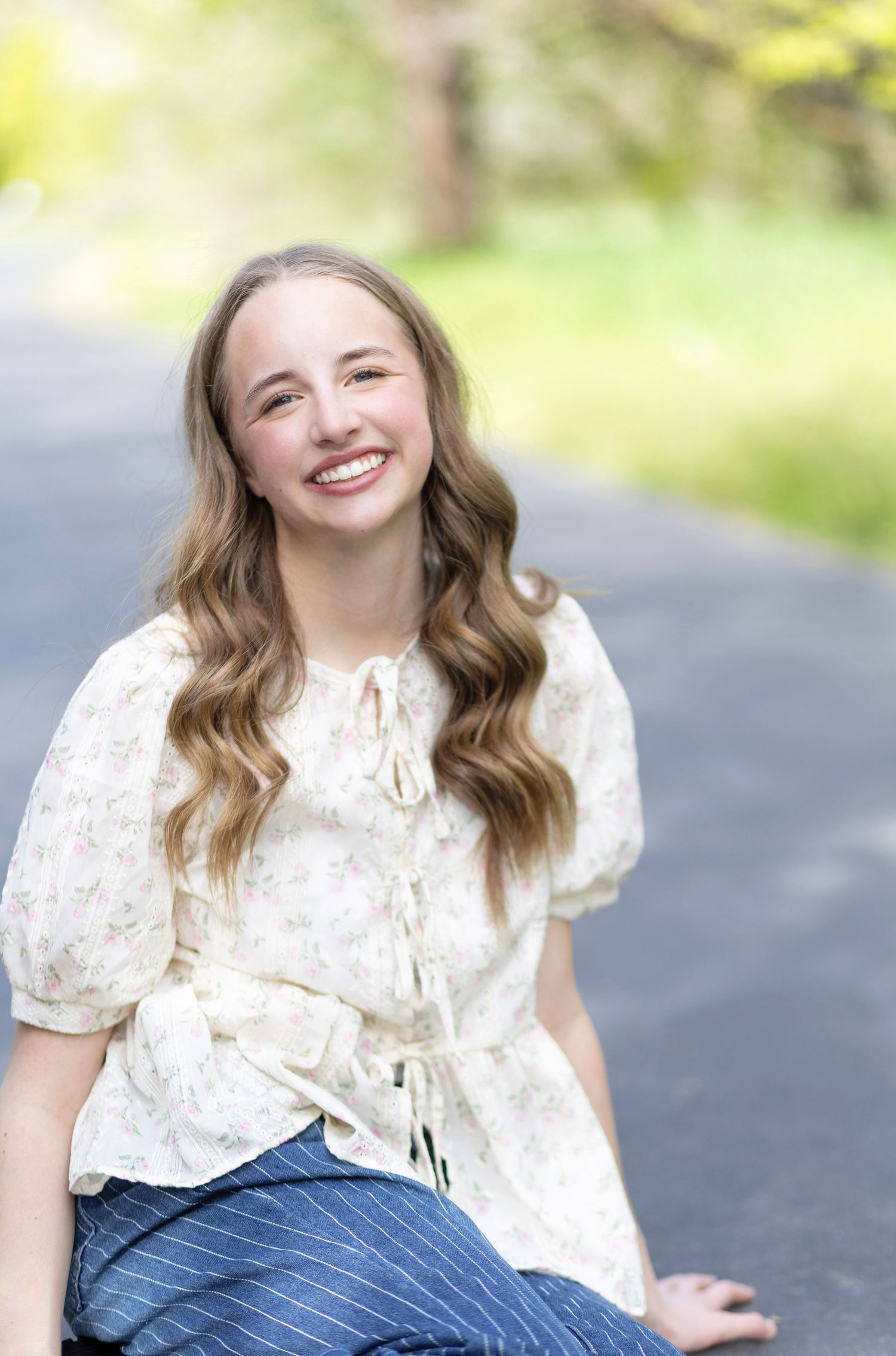 Smiling teenage girl with wavy blonde hair wearing a floral blouse and blue striped pants, sitting outdoors on a path.