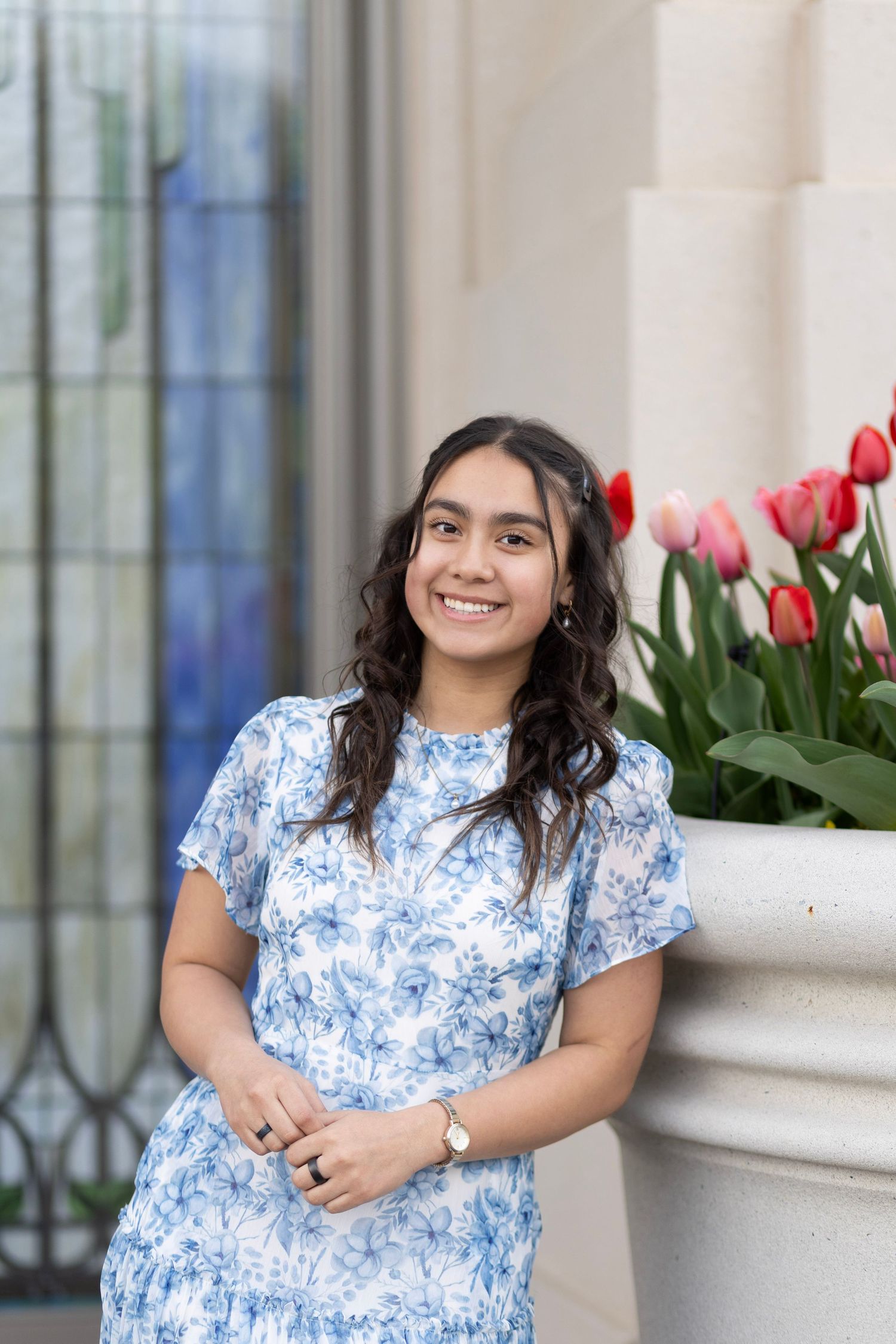 Young woman in blue floral dress smiling outdoors near pink tulips and a white column.