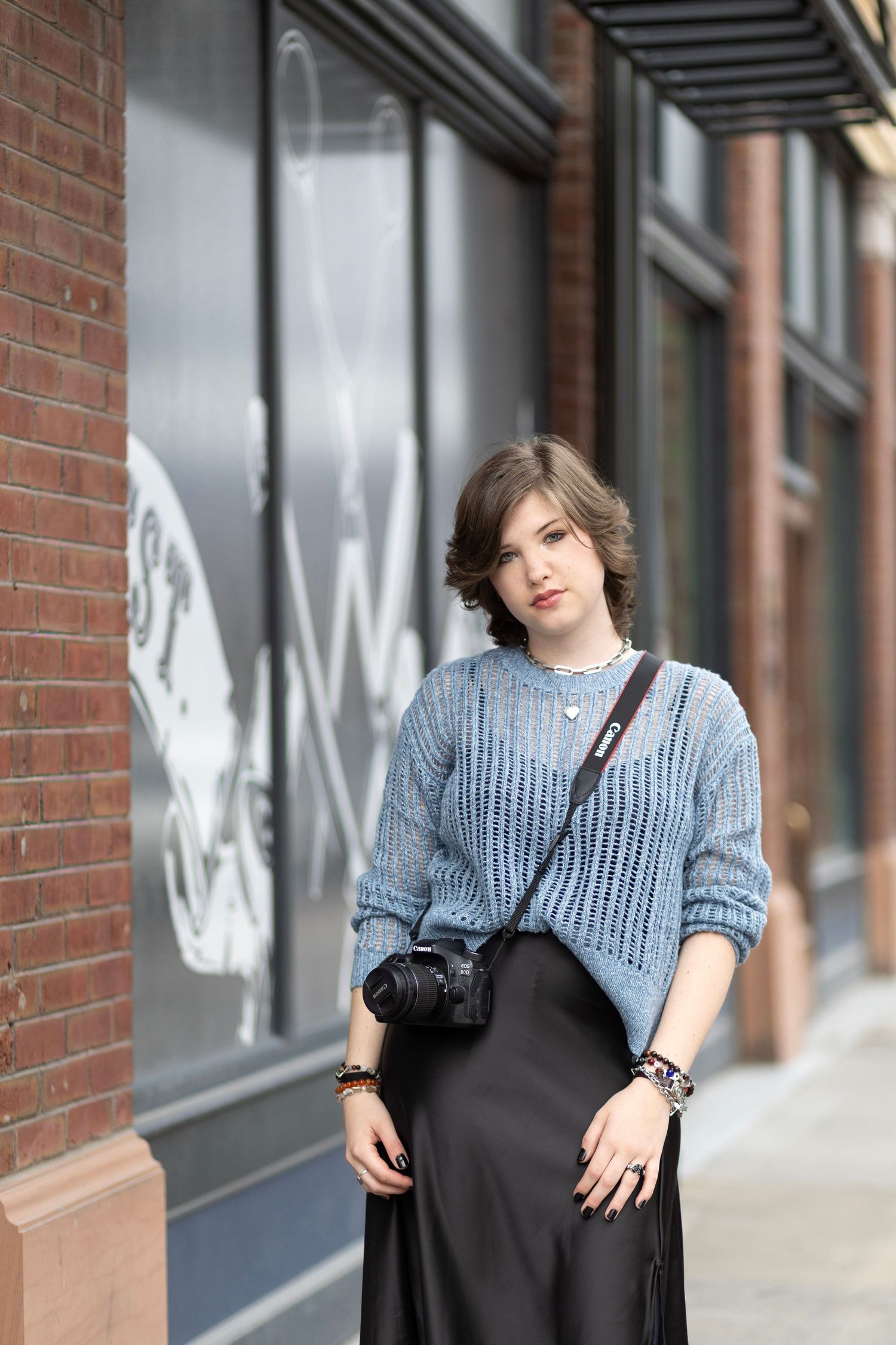 Young woman in blue knit sweater and black satin skirt poses on urban sidewalk with camera crossbody bag.