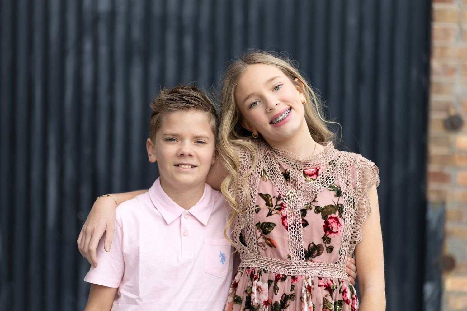 Two smiling children, a boy in pink polo and girl in floral dress, pose together outdoors near a dark wall.