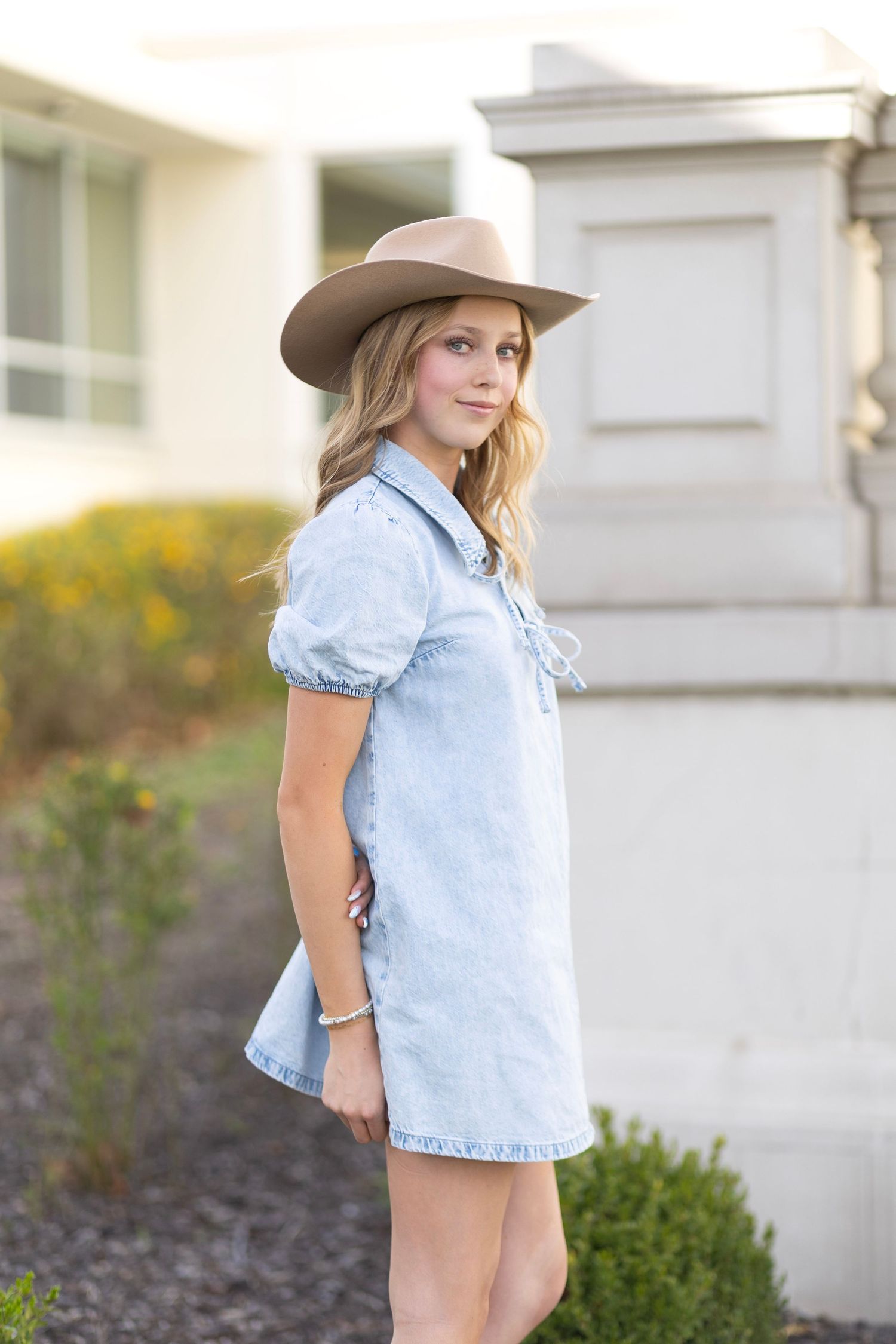 Young woman in a light denim dress and tan cowboy hat posing outdoors near greenery and a stone pillar.