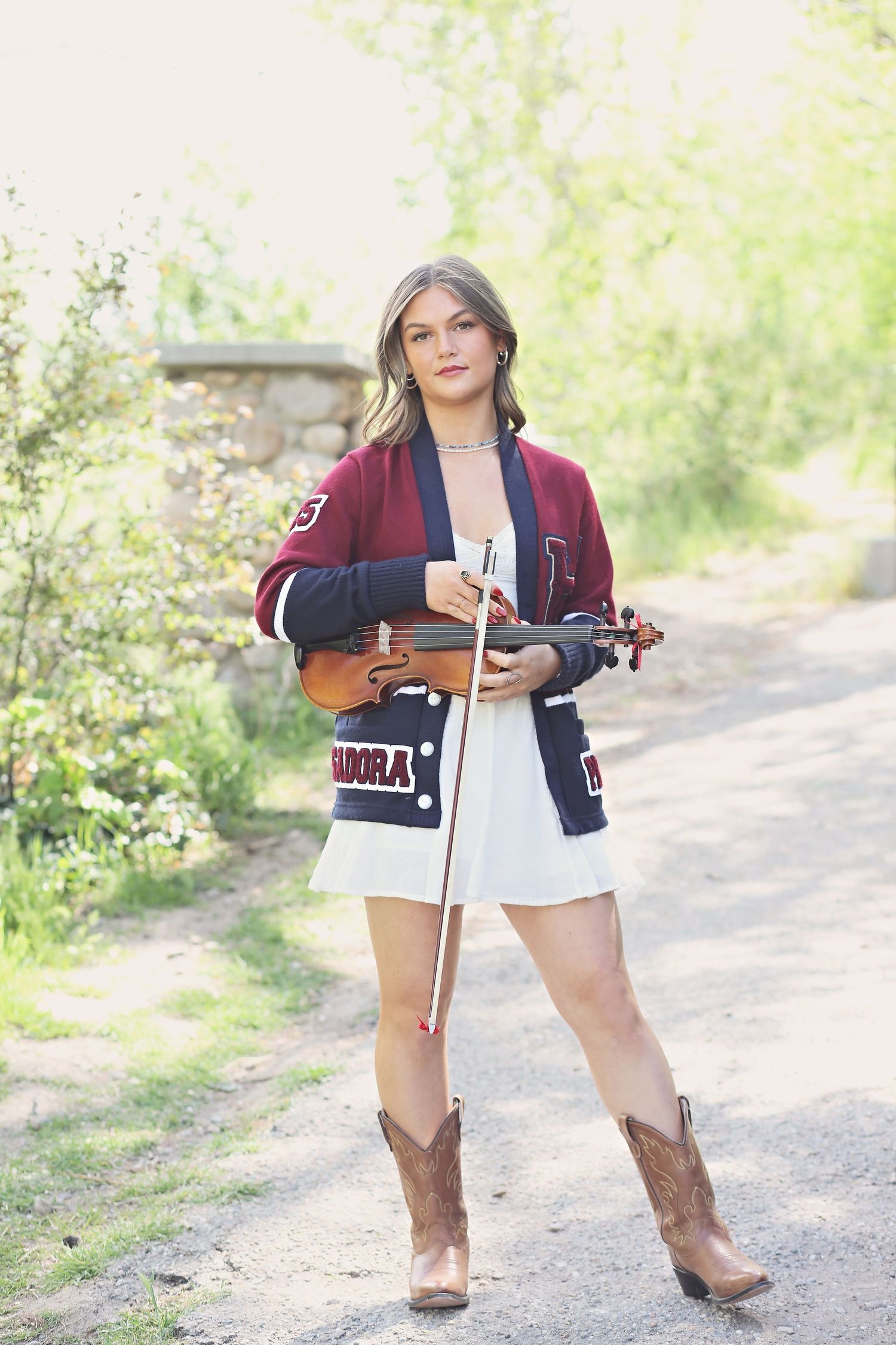 A woman in a burgundy varsity jacket and brown cowboy boots poses outdoors on a dirt path.