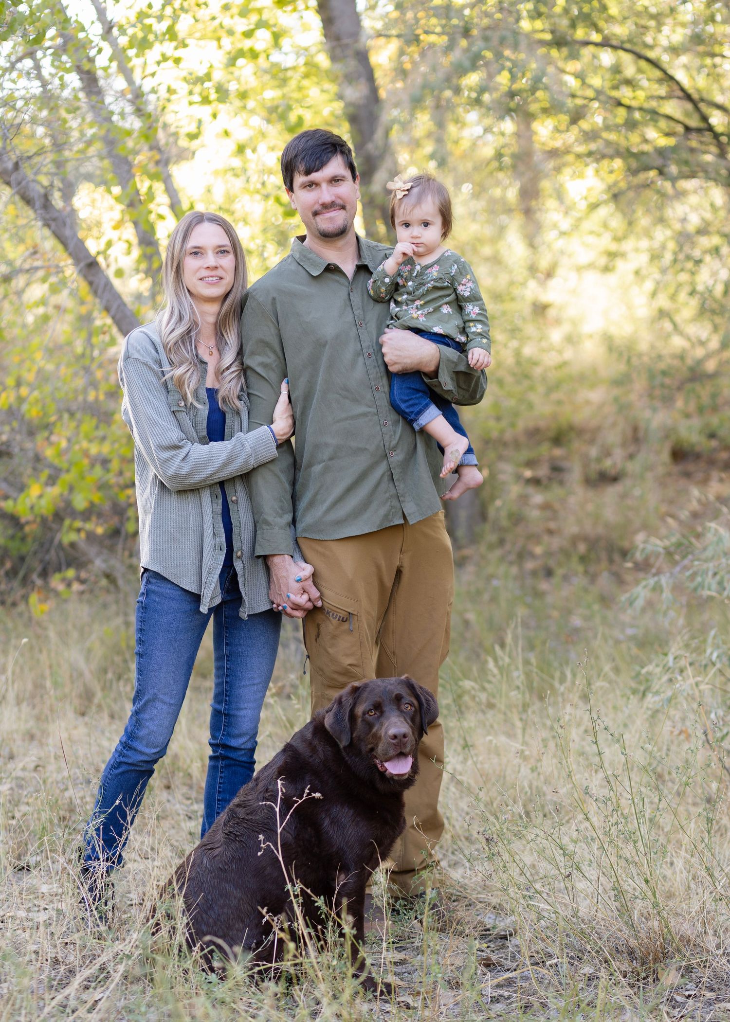 Family portrait with chocolate Labrador dog in natural outdoor setting with autumn foliage.