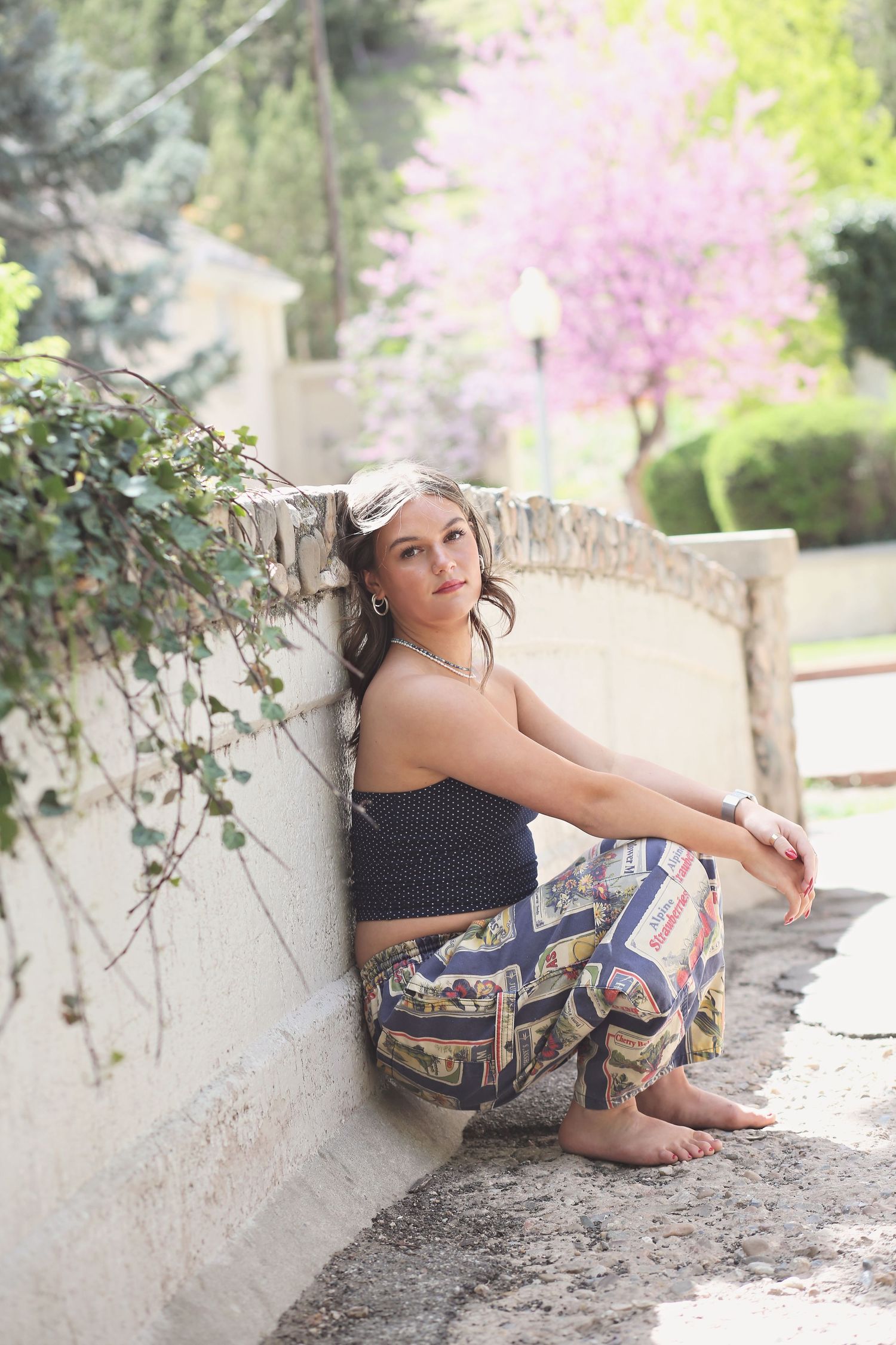 Fashion portrait of someone in a navy polka dot top and patterned pants lounging against a stone wall in a garden setting.