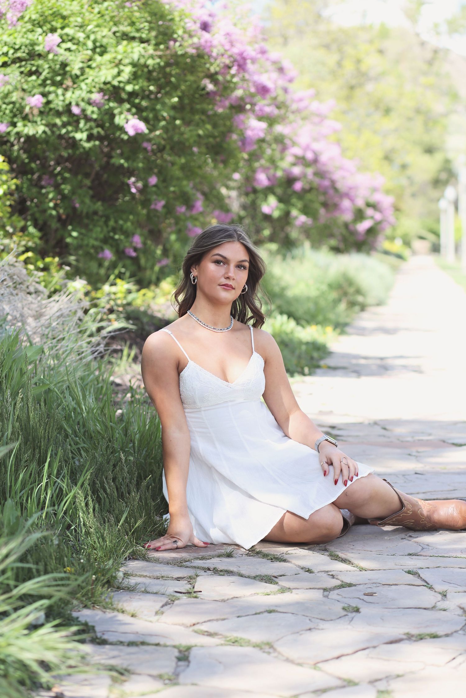 A figure in a white dress reclines on stone pavers surrounded by lush greenery and purple blooming flowers.