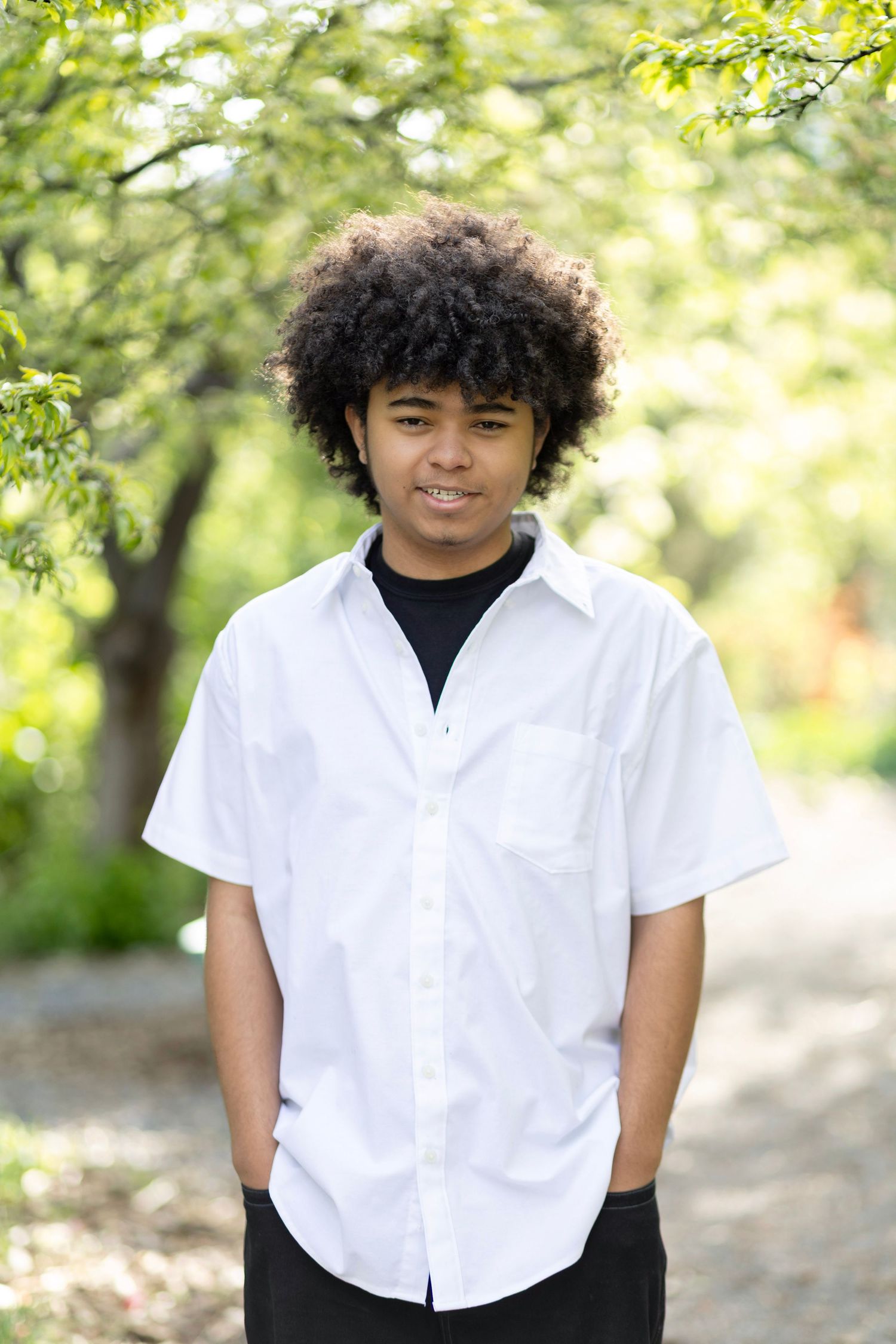 Young man with curly hair wearing a white button-up shirt, smiling outdoors in a green park setting.
