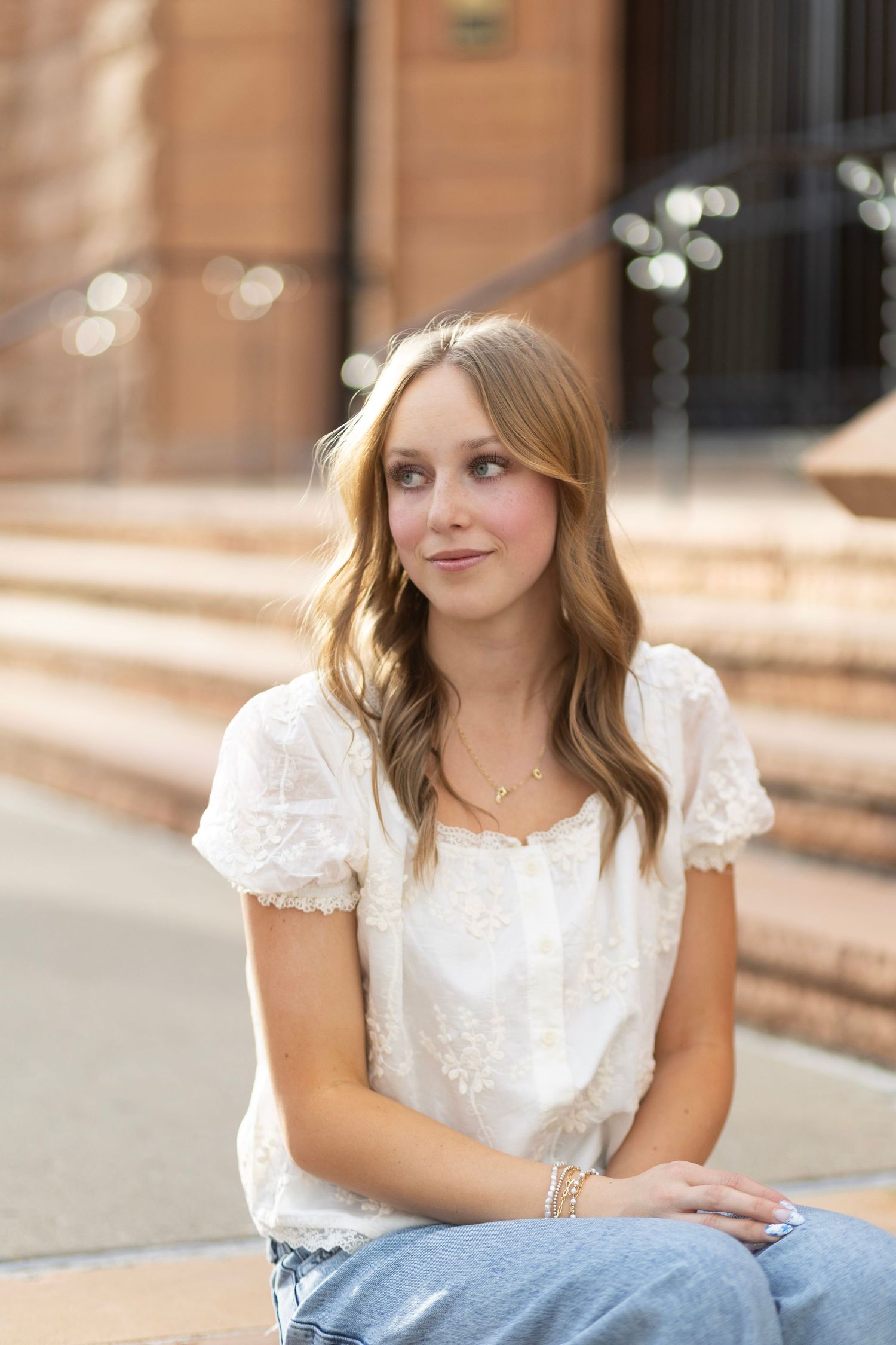 Young woman with wavy blonde hair wearing a white embroidered blouse and jeans, sitting outdoors on steps.
