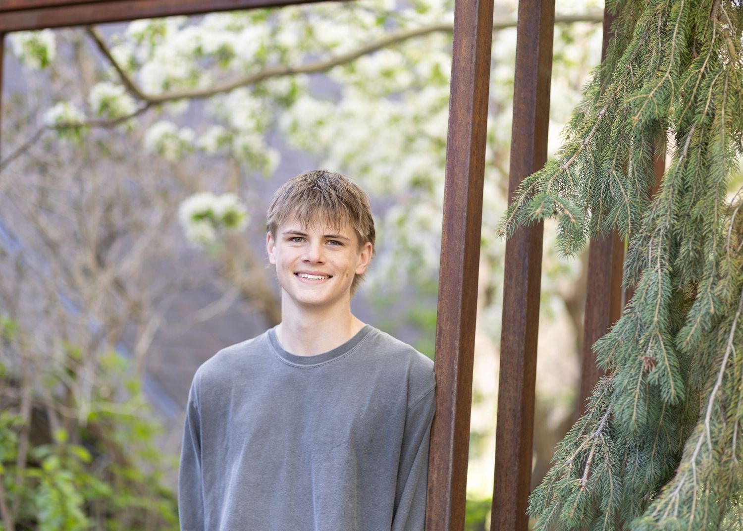 Smiling teenage boy in gray sweater standing outdoors near wooden frame and evergreen tree with spring blossoms.