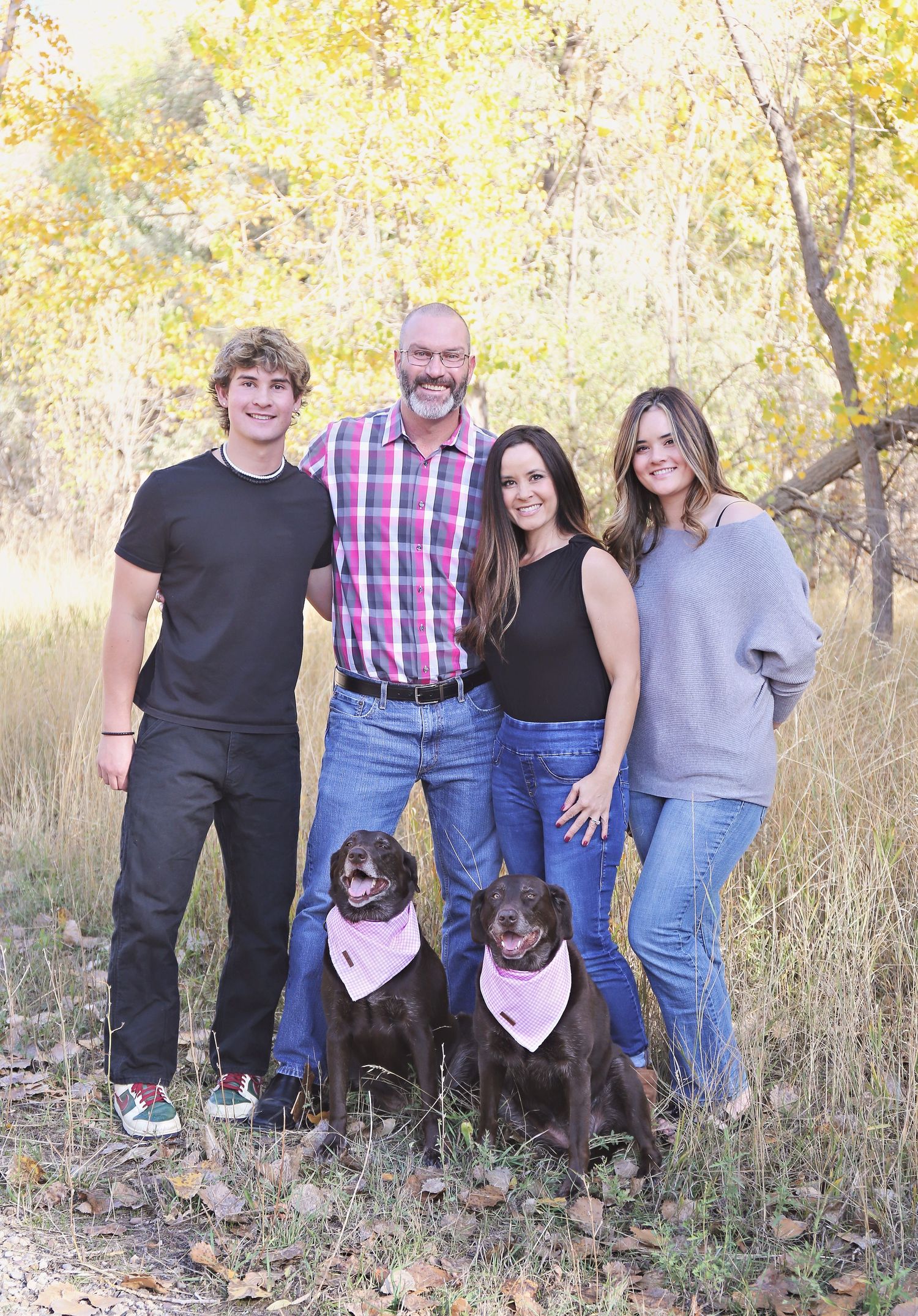 Extended family stands together in autumn field with golden grass and trees in background.
