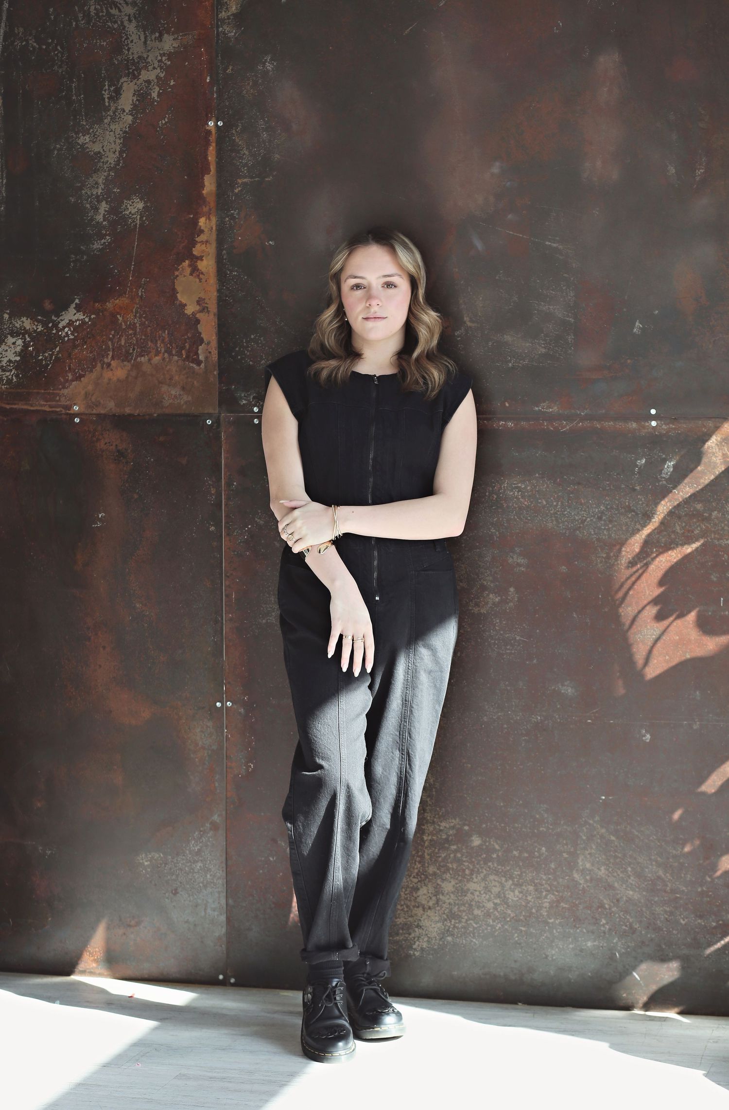 Person in black top and gray pants posing against textured metal wall with dramatic lighting.