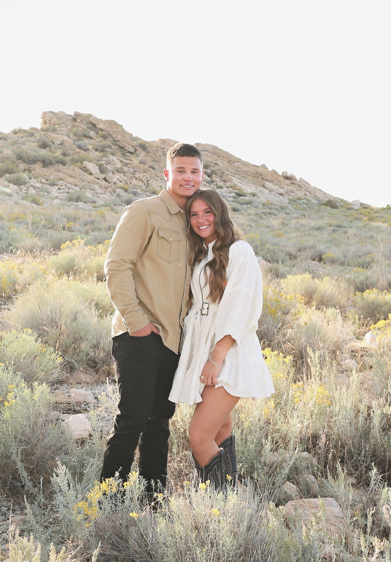 Couple standing close together in desert terrain with mountains and yellow wildflowers.