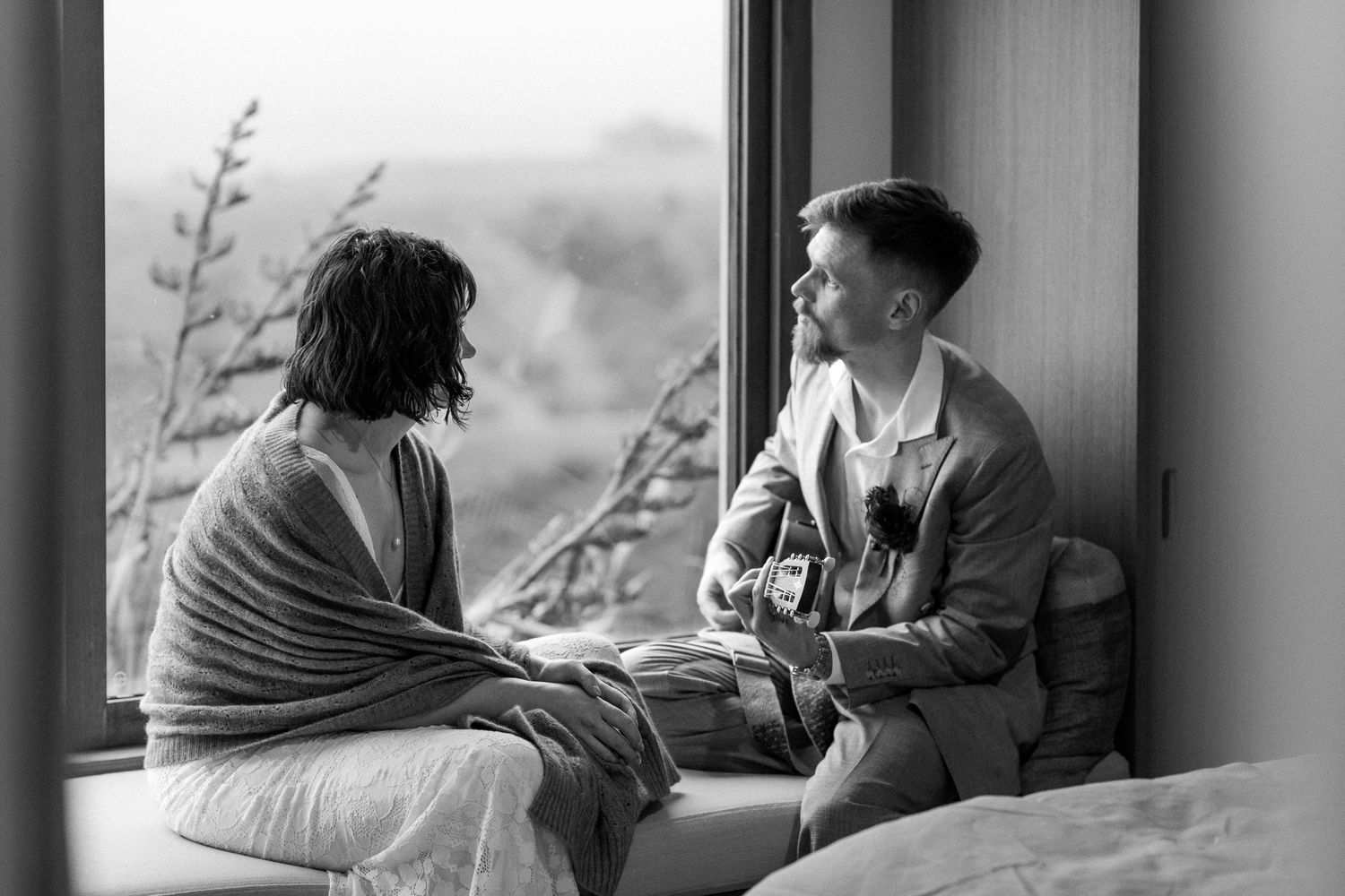 A black and white photo showing a groom playing guitar to his bride as they sit in a window box with a country view by August and Vine
