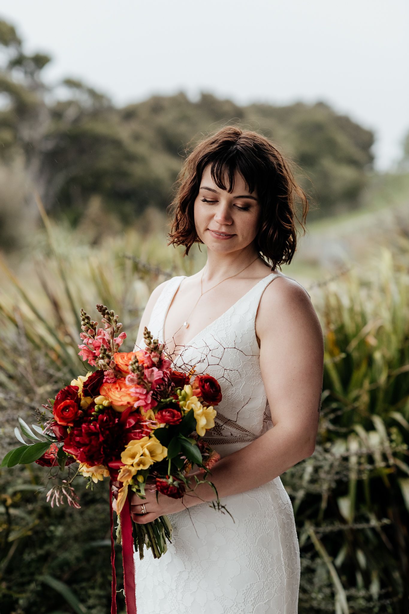 A bride in a white dress holds a vibrant autumn-colored bouquet with red and orange flowers against a natural backdrop by August and Vine
