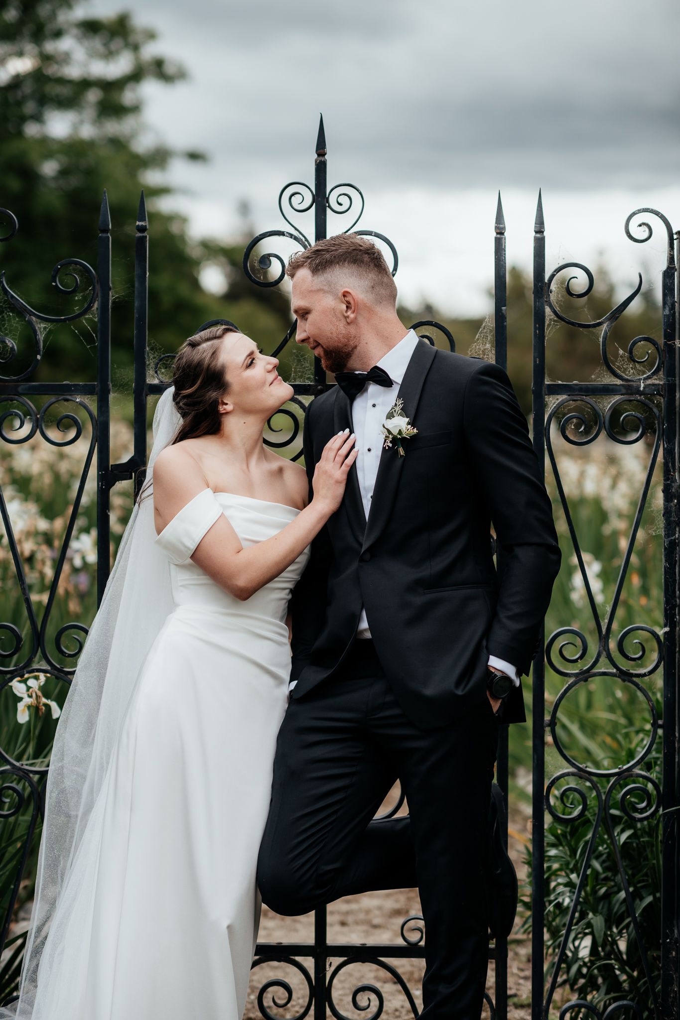 A couple in wedding attire share a romantic candid moment against an ornate metal garden gate at Lacewood Martinborough by August and Vine