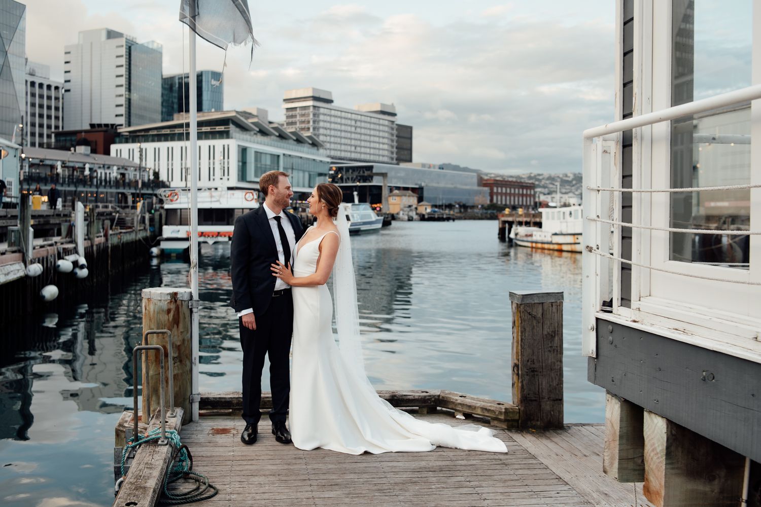 A wedding couple share a romantic moment on the dock at Dockside with boats and Wellington buildings in the background by August and Vine