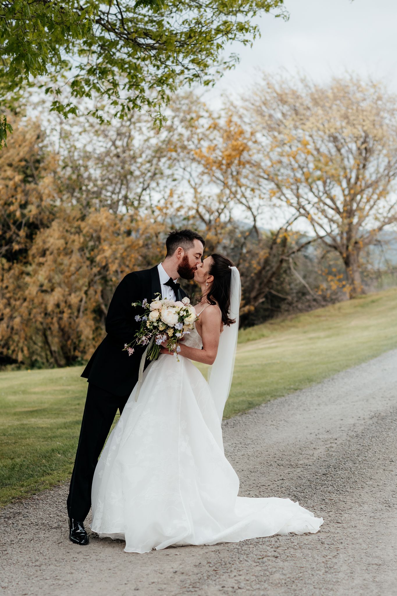 A romantic wedding couple share an intimate kiss on a gravel path surrounded by autumn trees at The Runholder by August and Vine