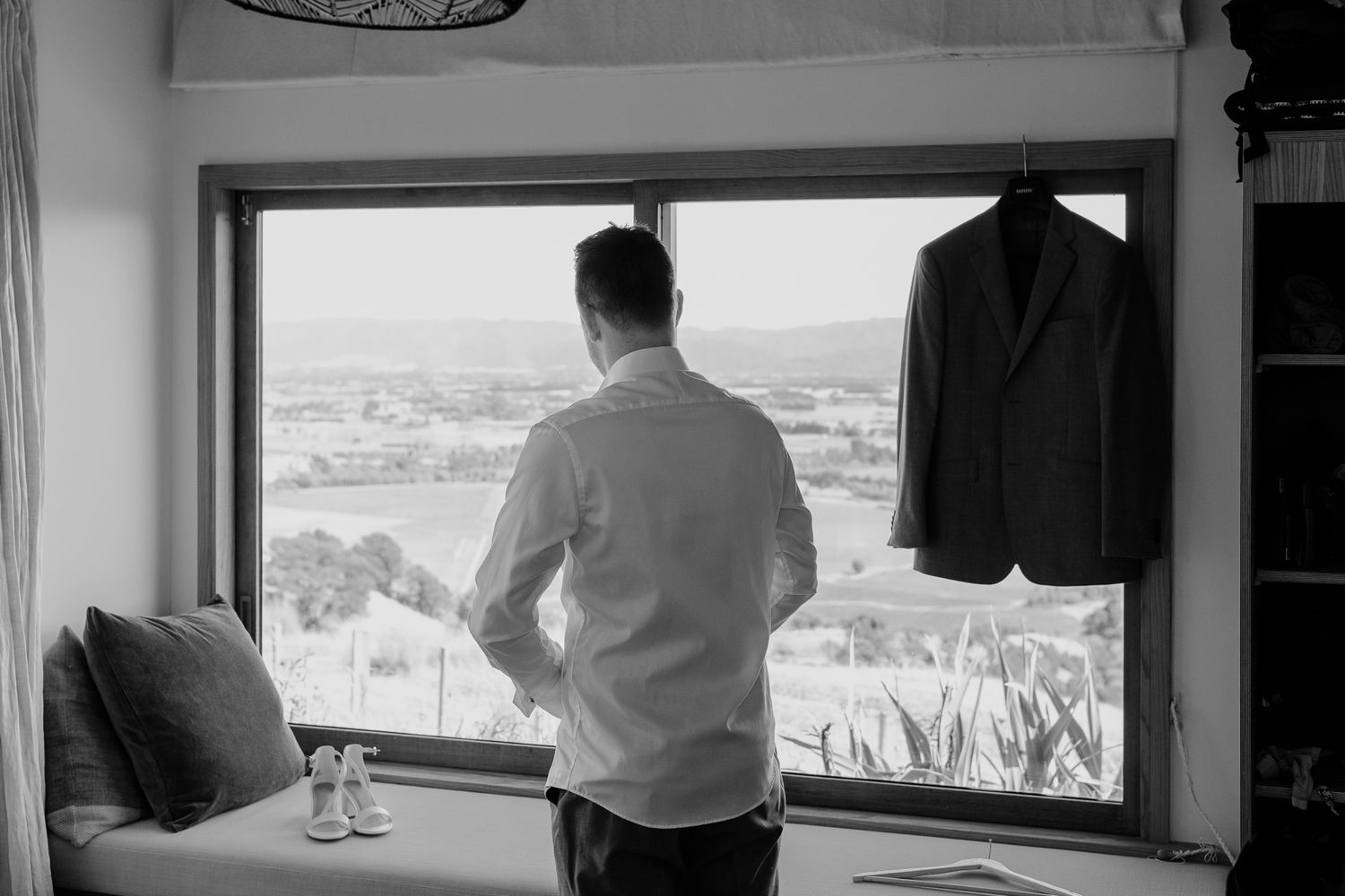 Groom stands at a window overlooking a scenic view, with a suit jacket hanging nearby at Whitimanuka by August and Vine