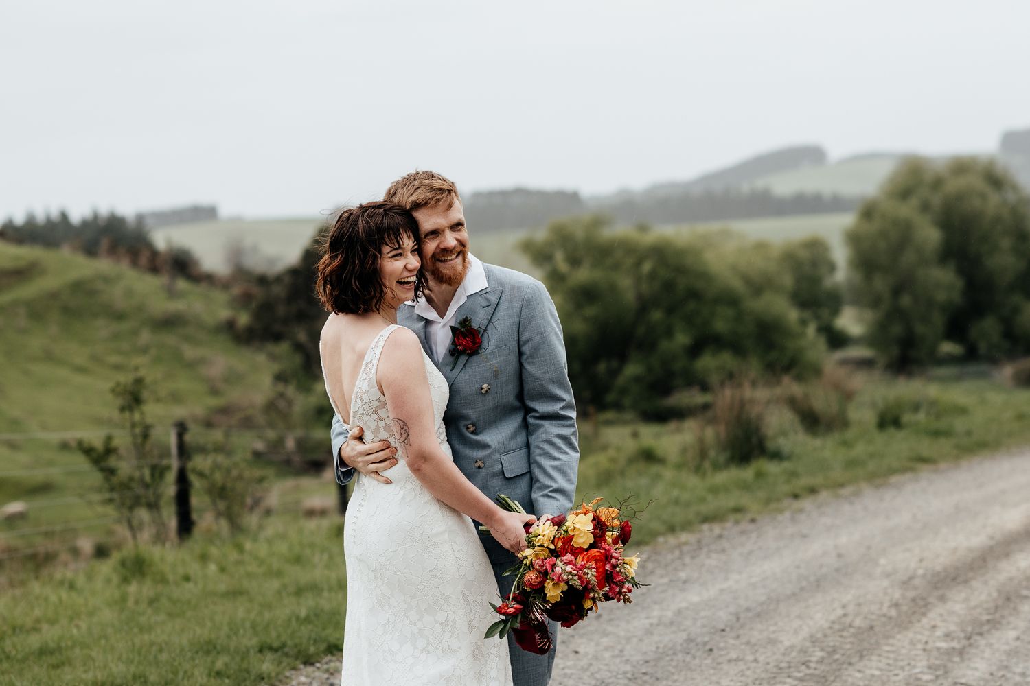 A couple in wedding attire share an intimate moment on a scenic country road surrounded by rolling green Wairarapa hills by August and Vine
