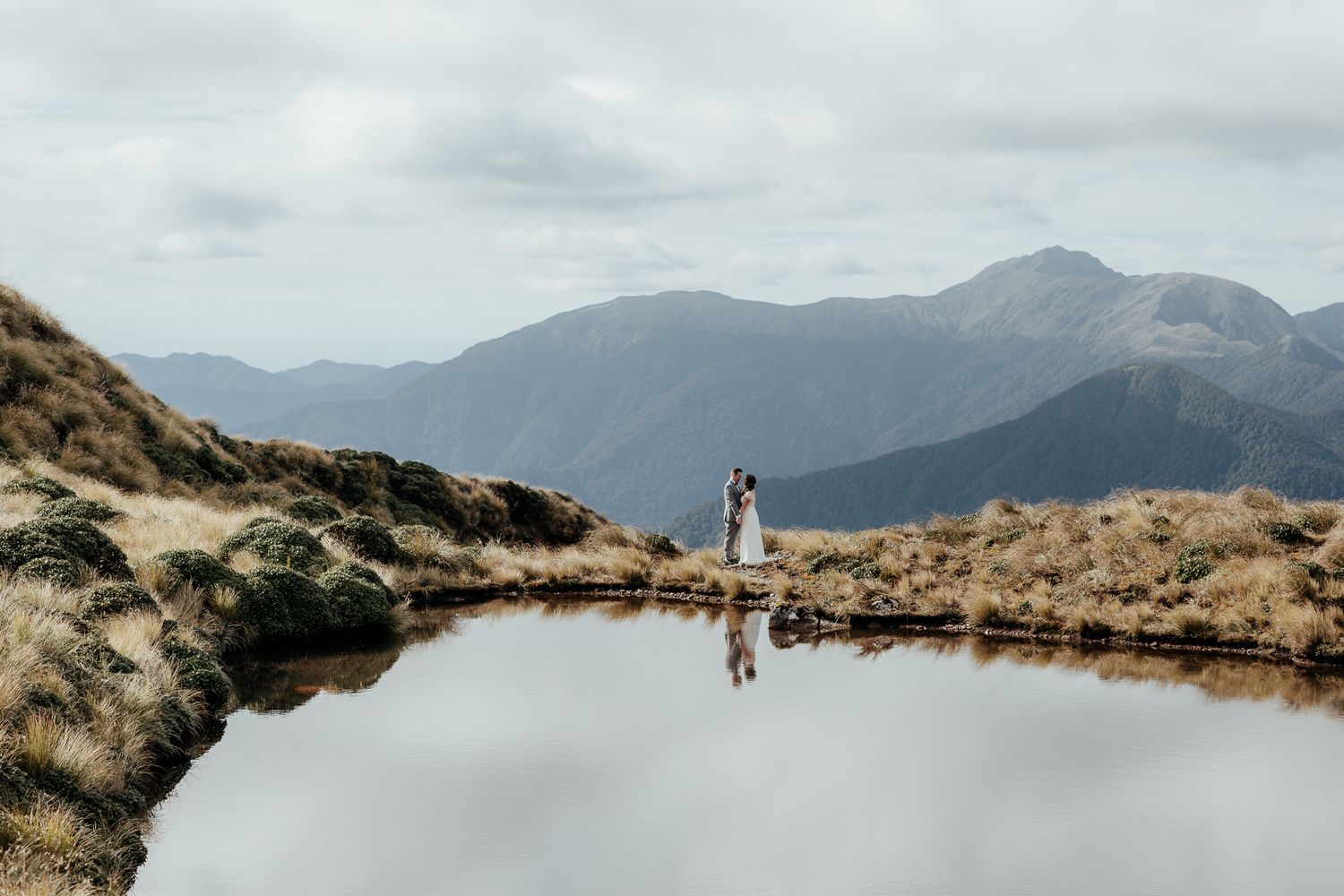 A wedding couple stand at an alpine lake in the Tararuas in Wairarapa with a scenic mountain landscape panorama behind by August and Vine