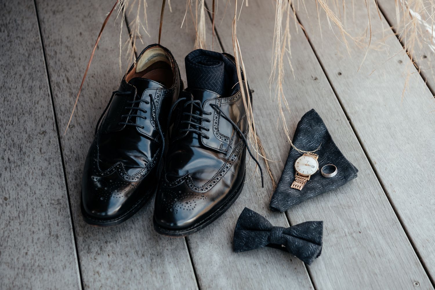 Shiny black formal dress shoes with bow tie and watch accessories arranged on wooden floor by August and Vine