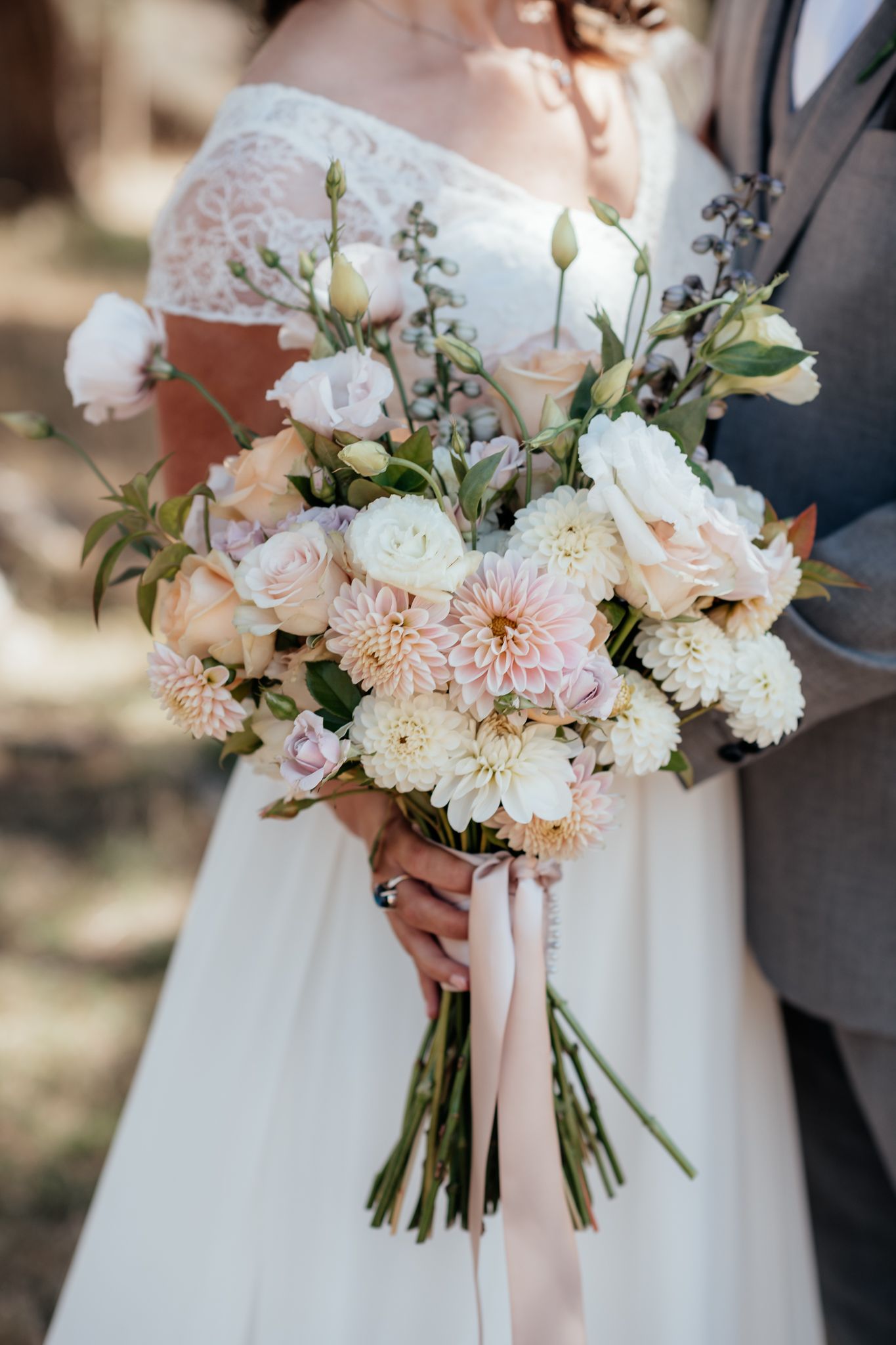 A romantic wedding bouquet featuring white roses, blush dahlias, and delicate greenery tied with pink silk ribbon captured by August & Vine