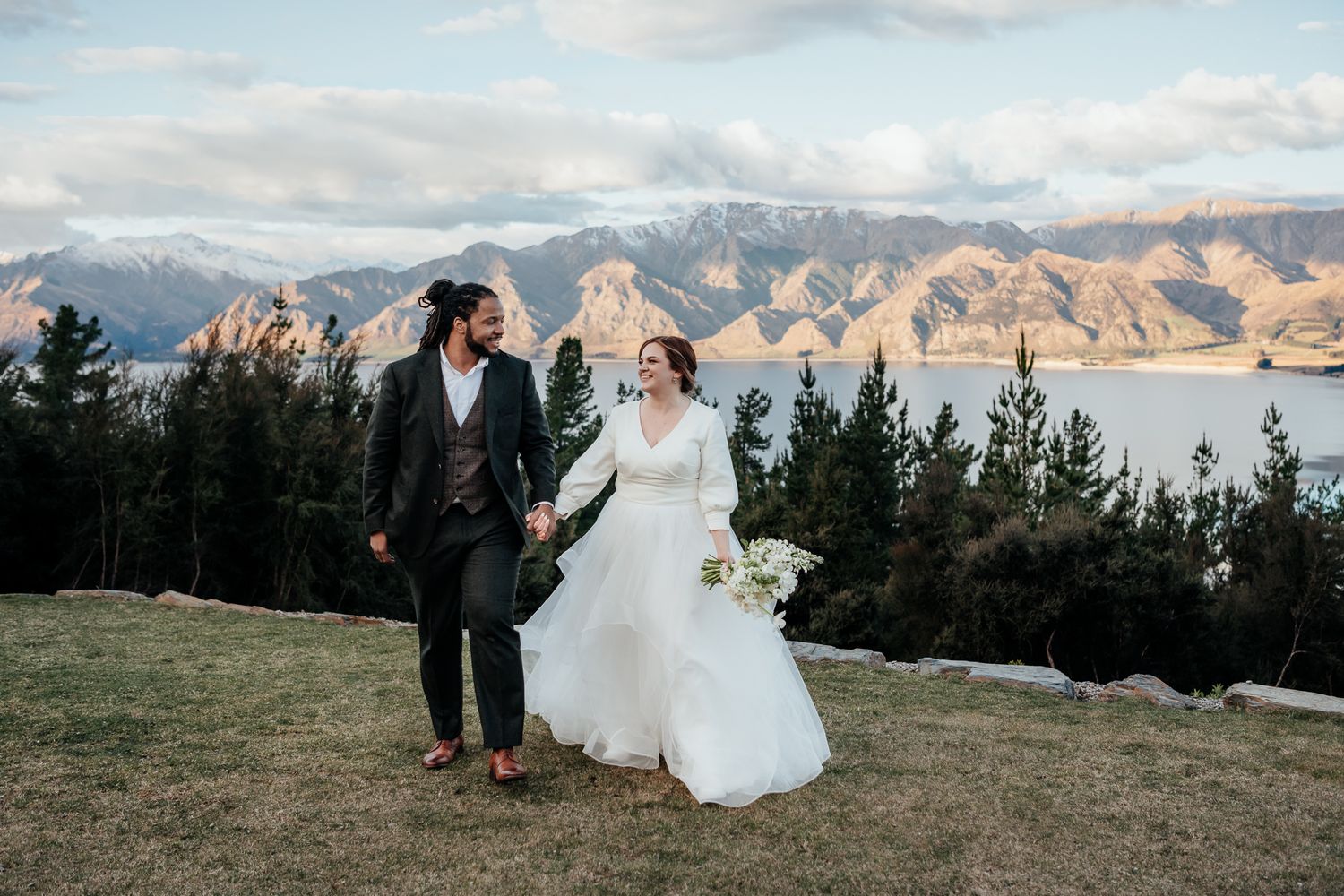 Couple in wedding attire walk hand in hand with scenic mountain views behind at Lake Hawea View during sunset by August and Vine
