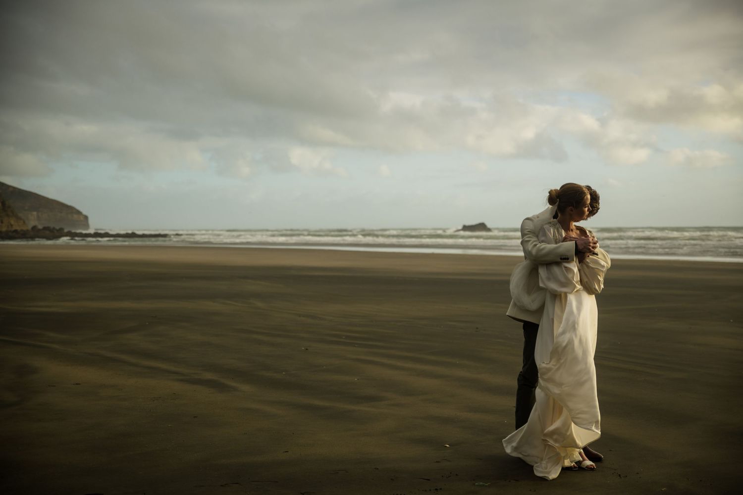 Bride and groom embrace on a misty NZ beach at sunset with waves crashing in the background by August and Vine