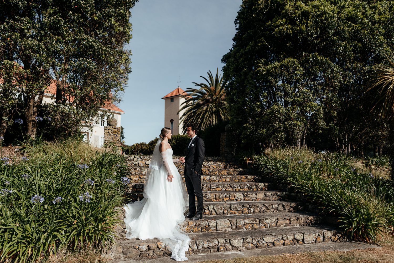A couple in wedding attire stand on stone steps surrounded by lush gardens and palm trees at historic Rototawai by August and Vine