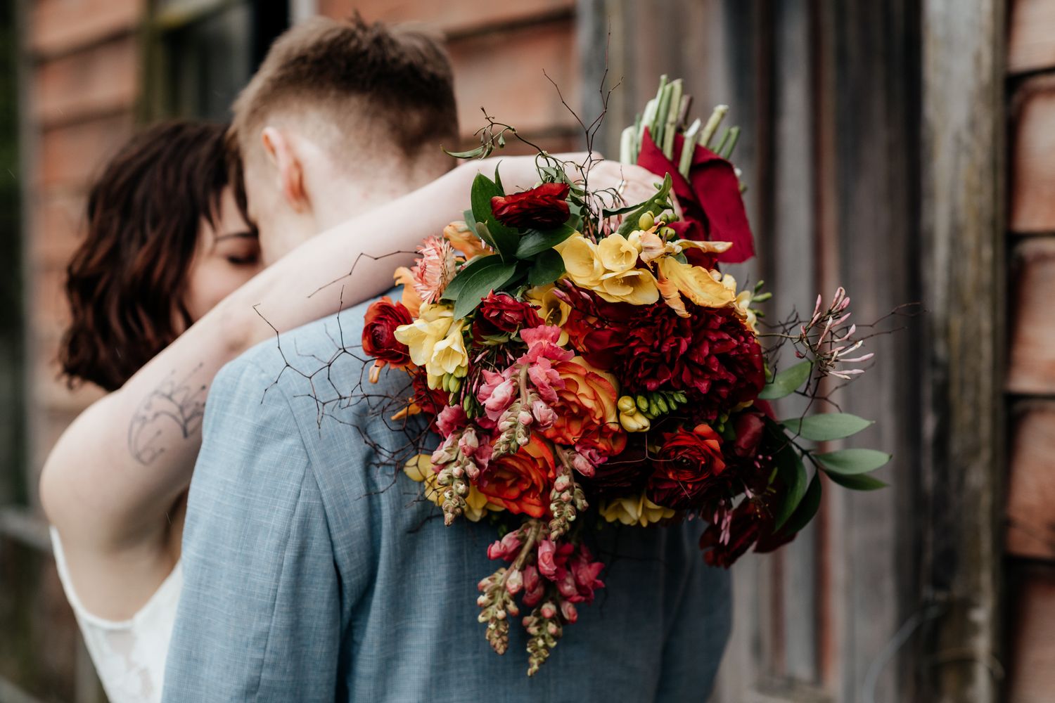 A bride and groom embrace during a romantic autumn Wairarapa wedding with vibrant red and orange floral bouquet by August and Vine