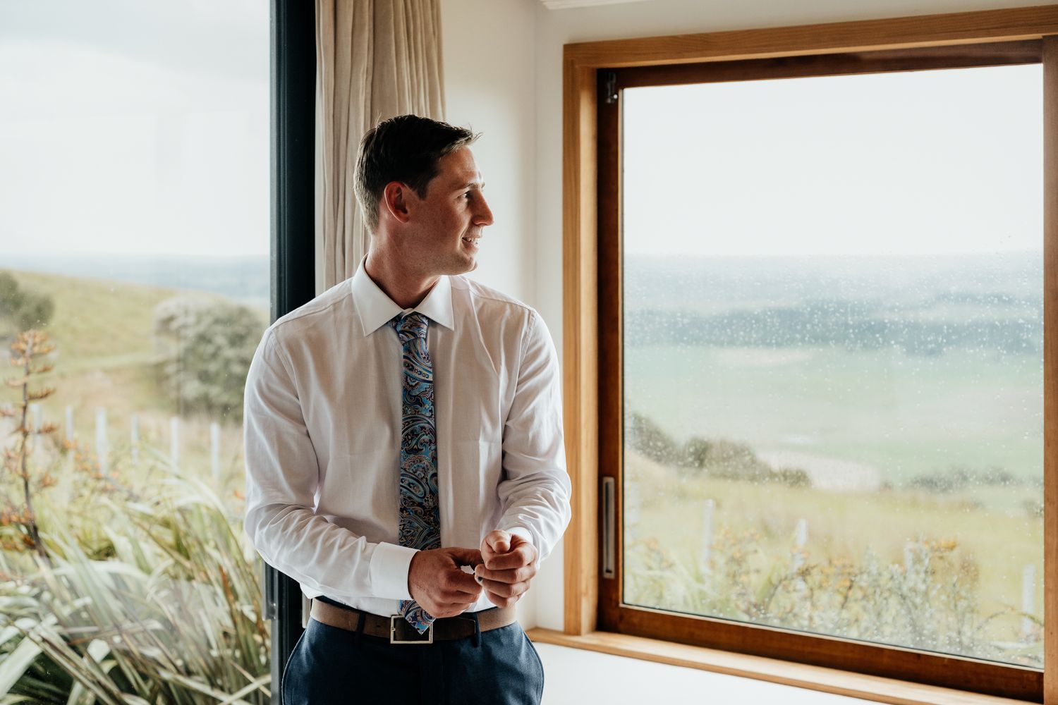 Groom in white shirt looking out large window at scenic mountain landscape view in Wairarapa by August and Vine