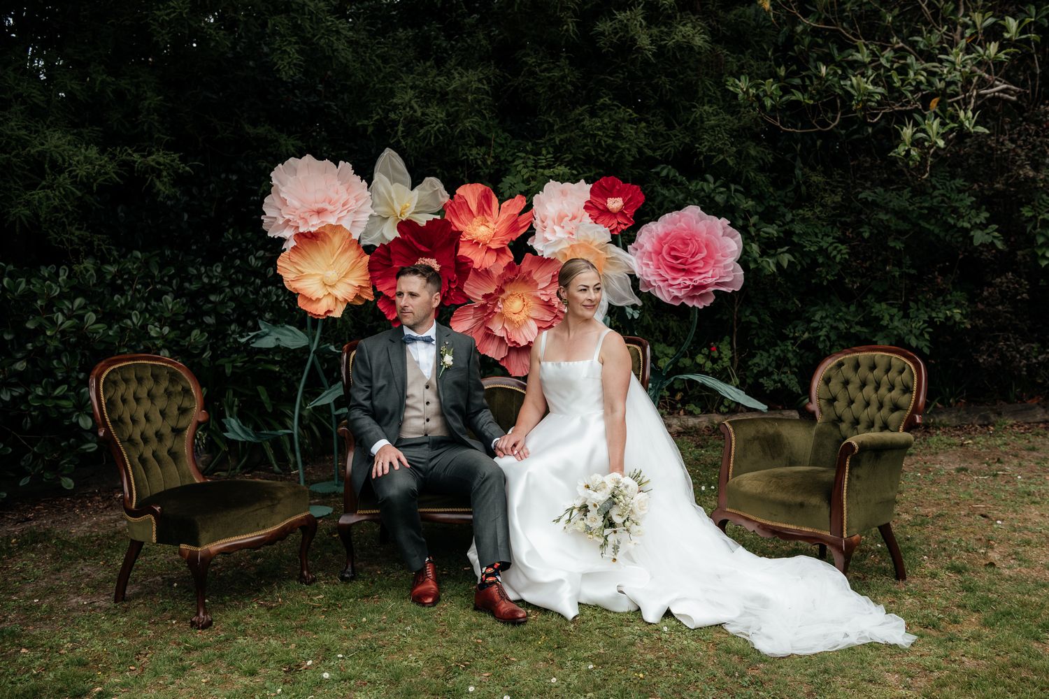 A wedding couple sit together on vintage chairs in the garden at Tarureka with giant, bright paper flowers behind them by August and Vine