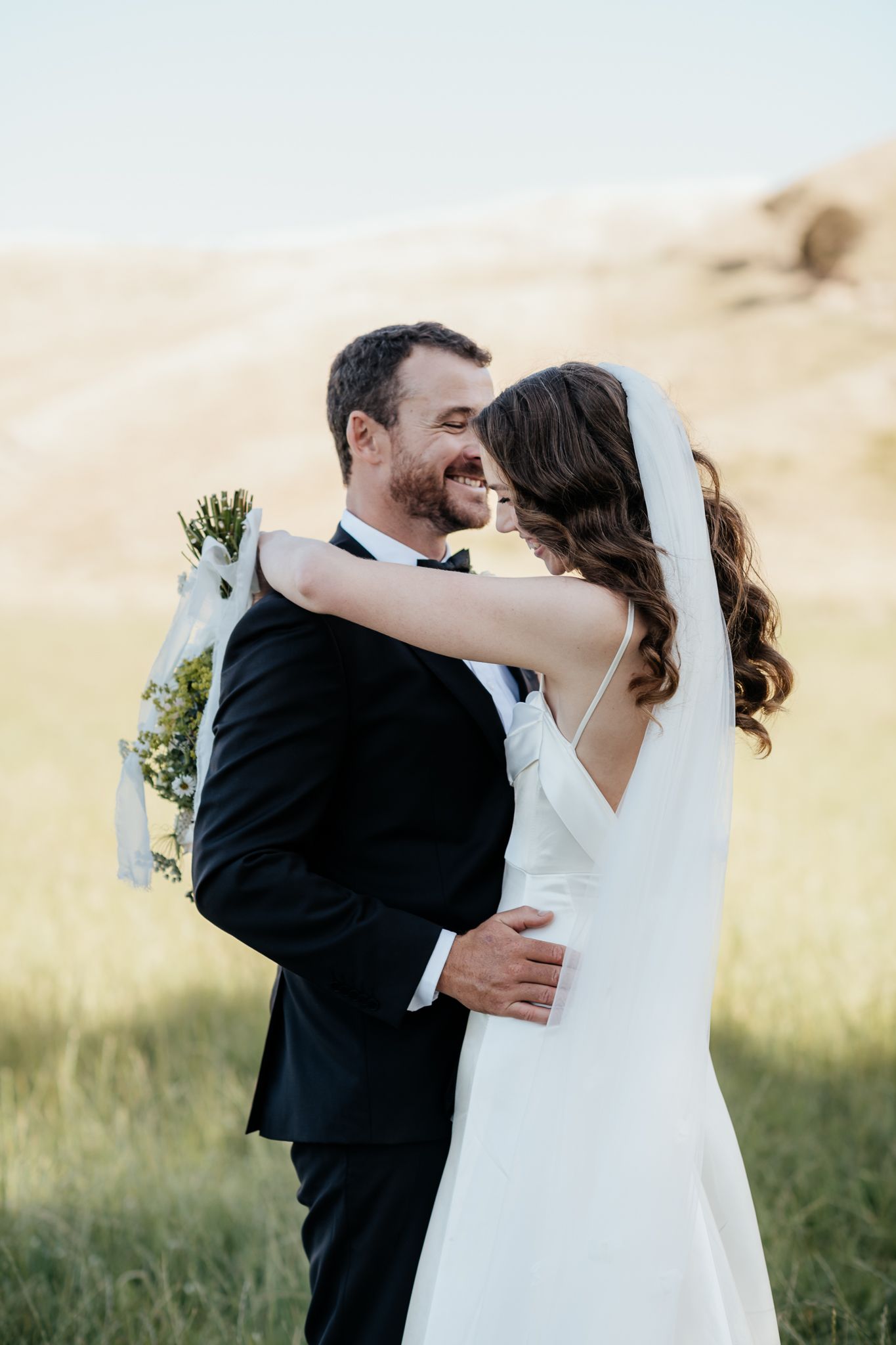 Elegant Bride and Groom in black tie embrace laughing on a farm at Riversdale captured by August and Vine