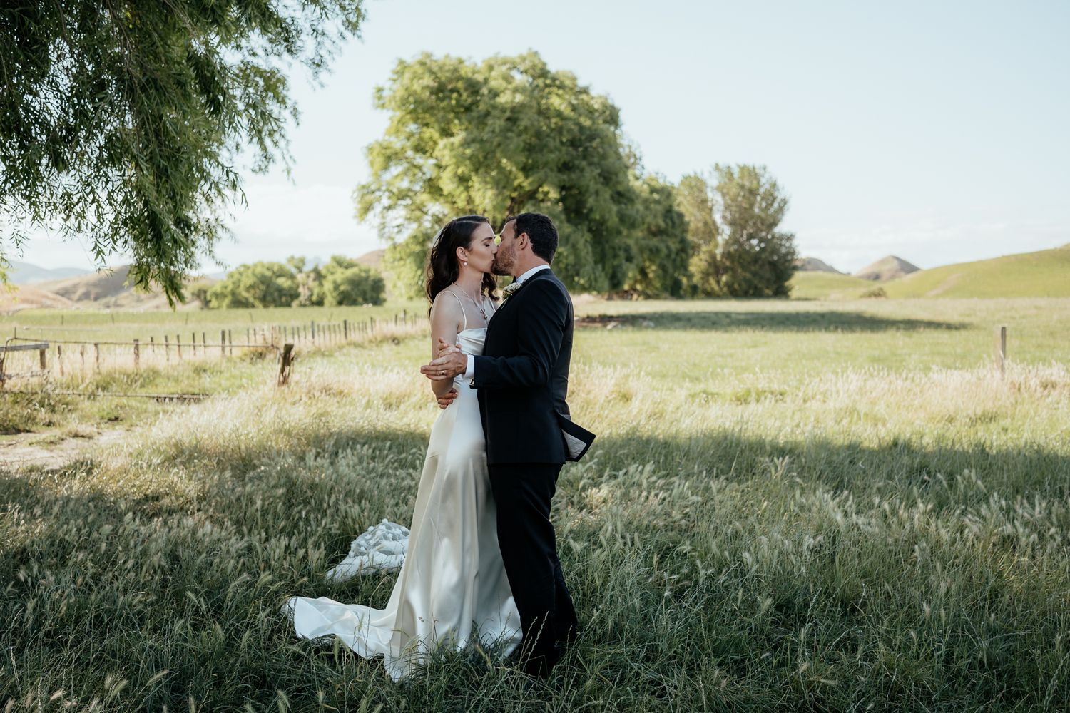 Couple embraces in a romantic moment surrounded by tall grass and natural scenery at golden hour in Wairarapa by August and Vine