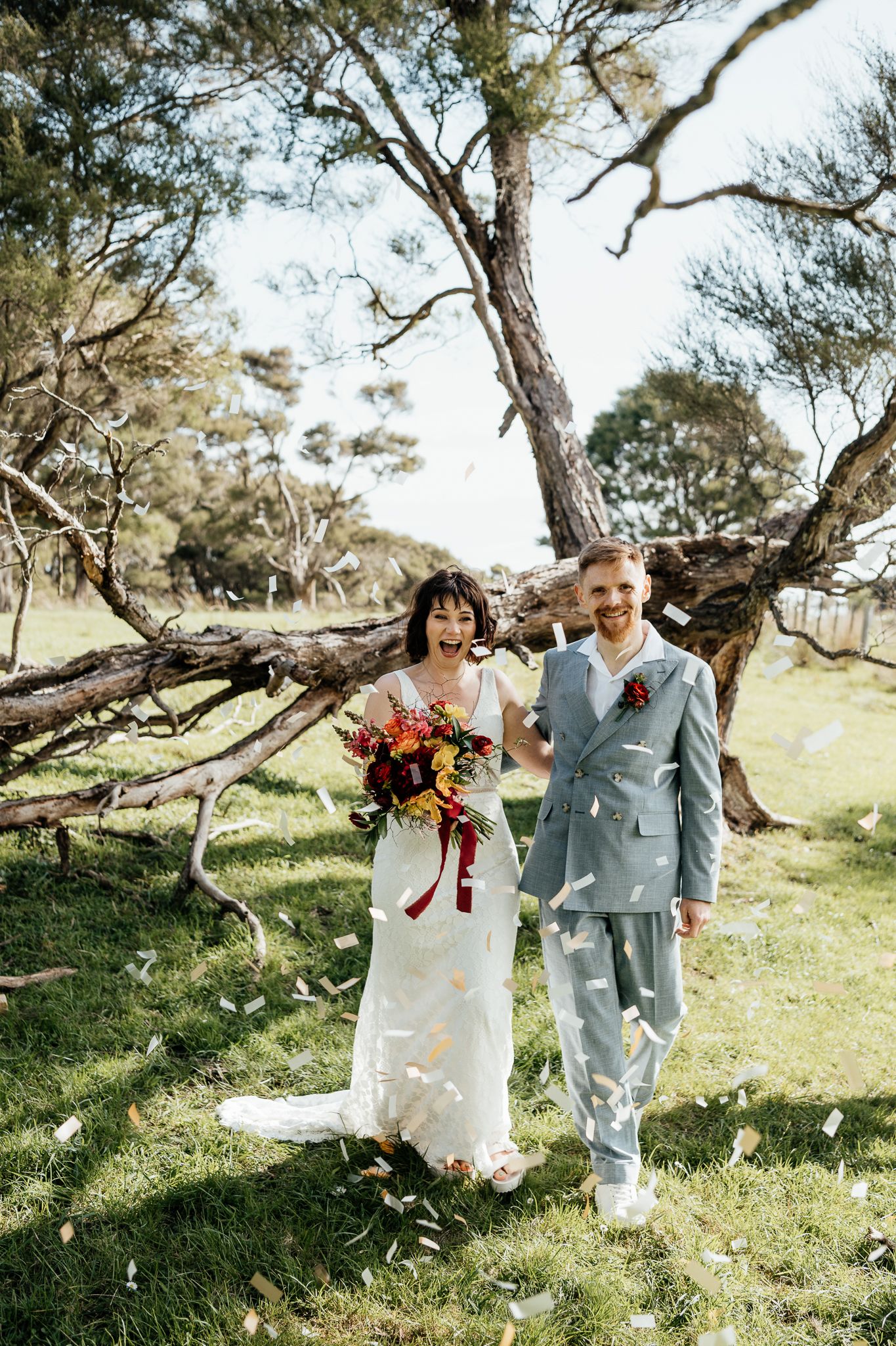 A wedding couple are showered in confetti under a tree at Whitimanuka Wairarapa with the bride holding a vibrant bouquet by August and Vine