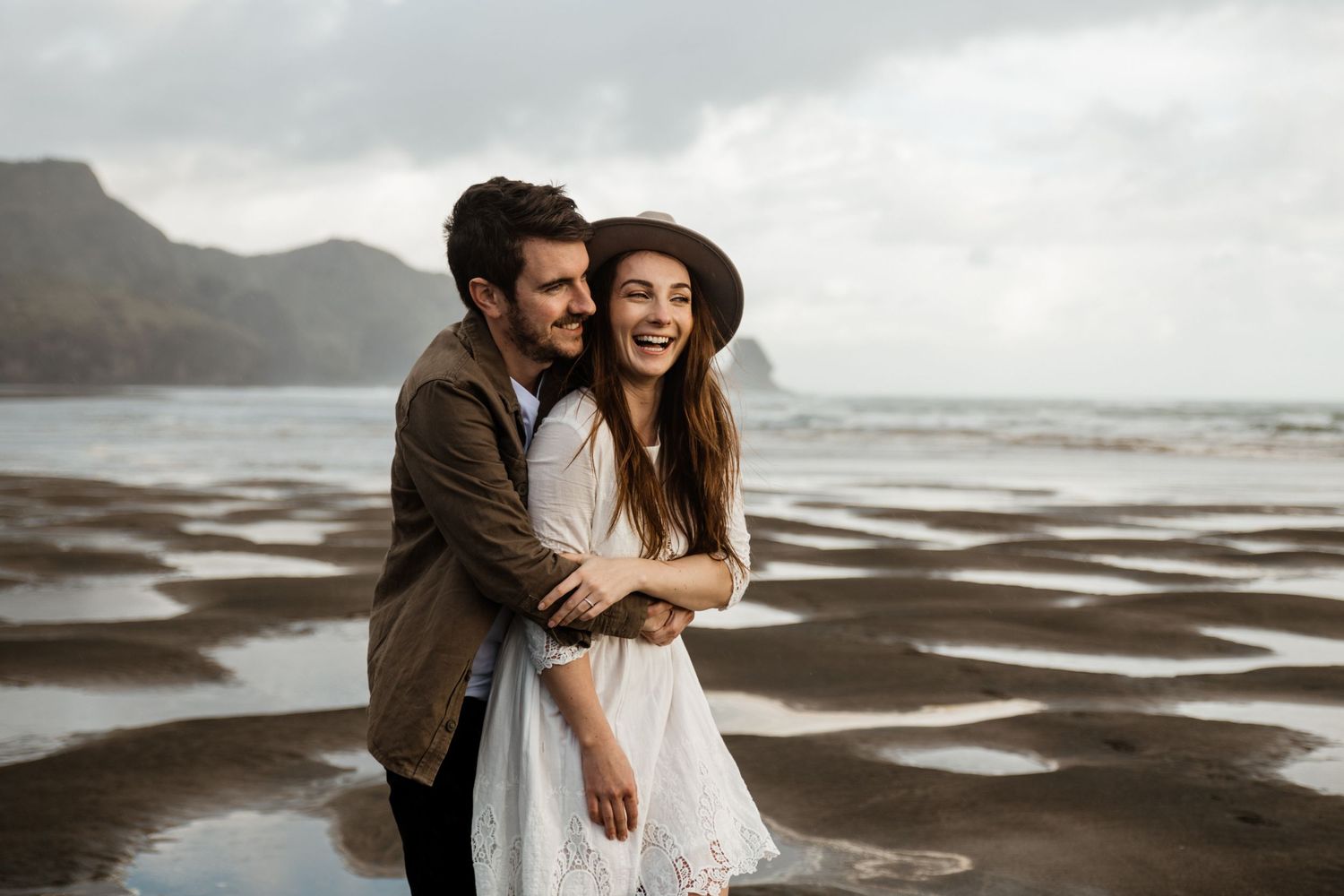 An engaged couple share an intimate moment on a moody beach with dramatic cloudy skies in the background by August and Vine Photography