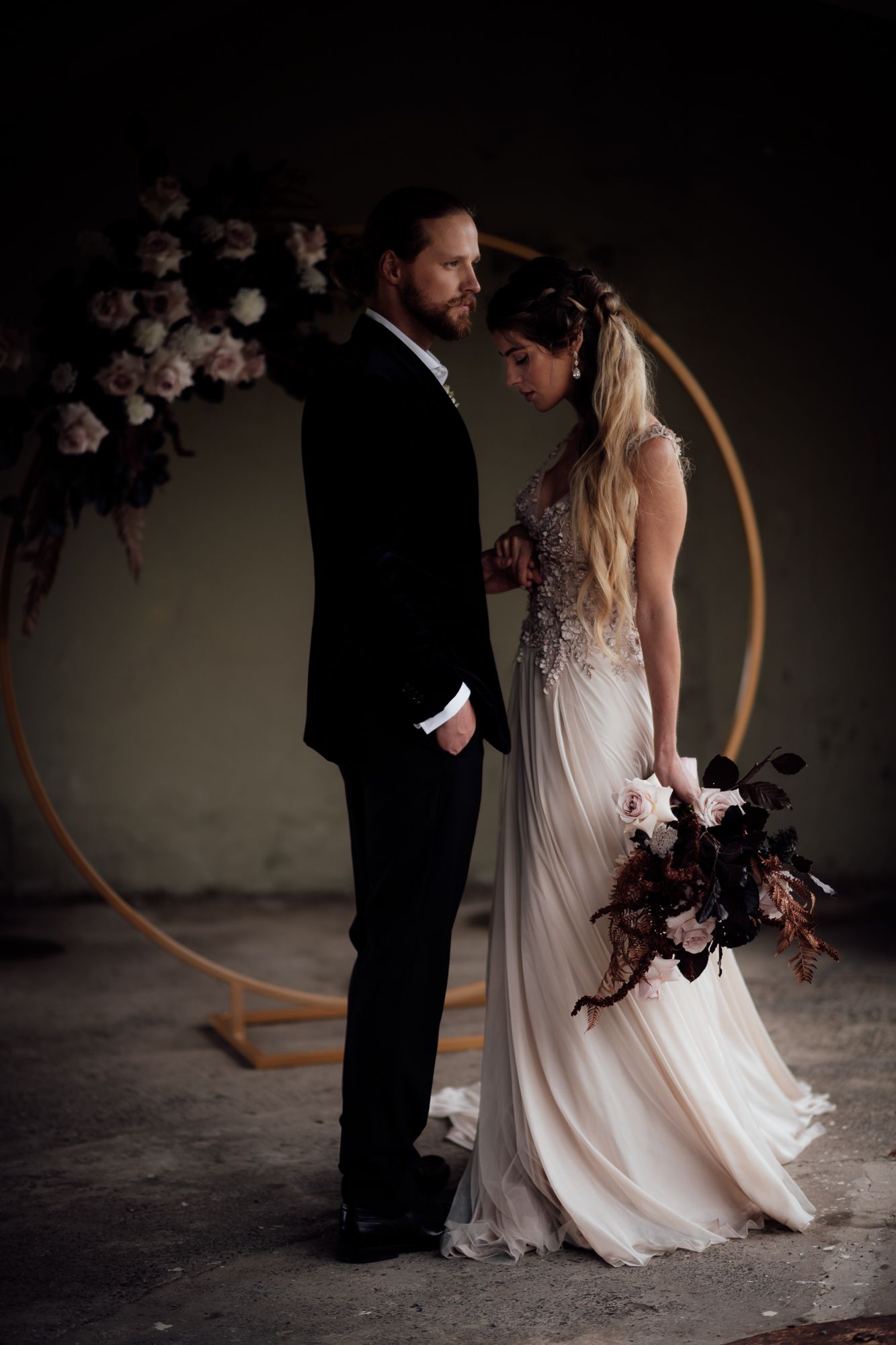 A wedding couple stand in a moody industrial setting with a large golden hoop backdrop with blush and black florals by August and Vine