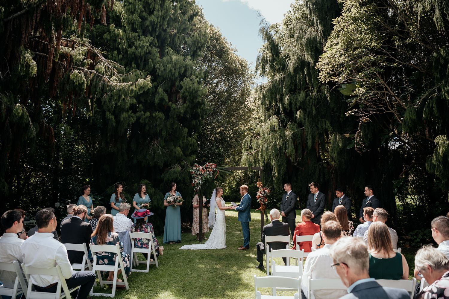 A beautiful outdoor wedding ceremony with white chairs in rows beneath tall trees and natural greenery at Anam Cara by August and Vine