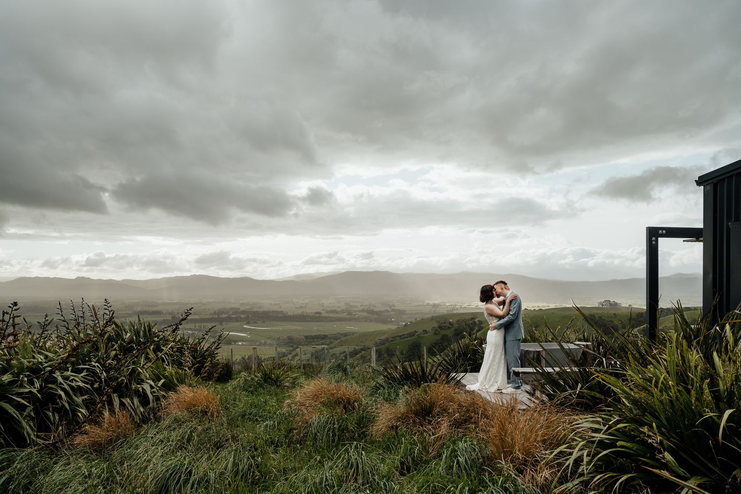 A scenic mountain landscape with dramatic cloudy skies stretches across a panoramic view of fields at Whitimanuka by August and Vine